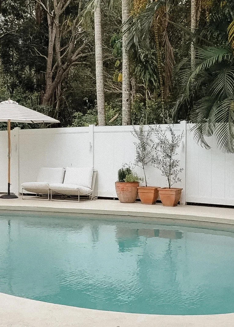 A backyard pool area with a white fence, potted plants, white outdoor sofa, and a patio umbrella, surrounding a swimming pool with calm water.