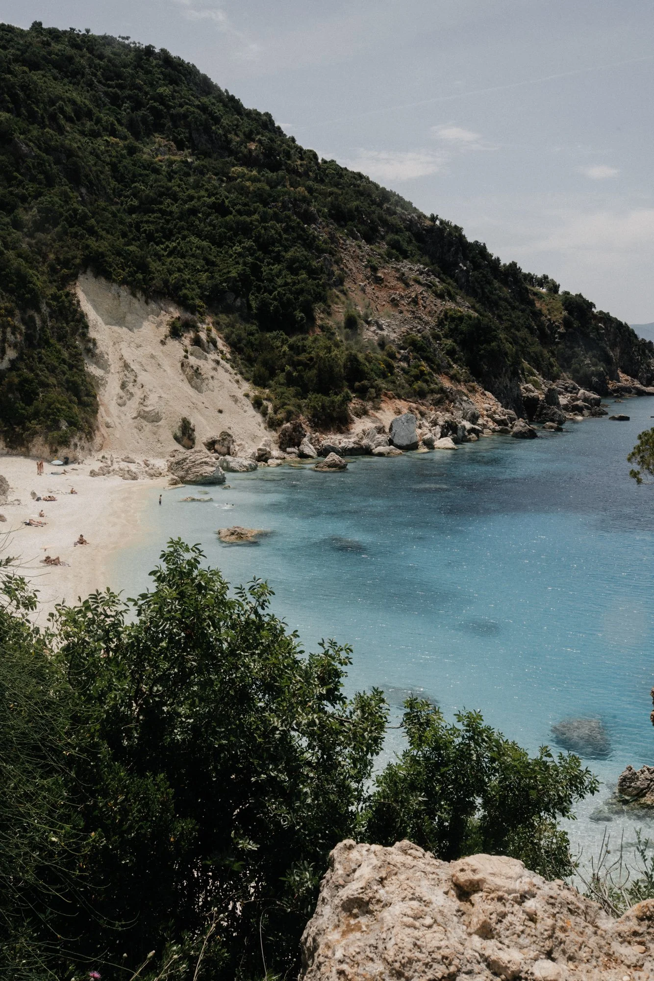 A scenic view of a coastal cove with clear blue water, a sandy beach with a few people, and lush green hills in the background.
