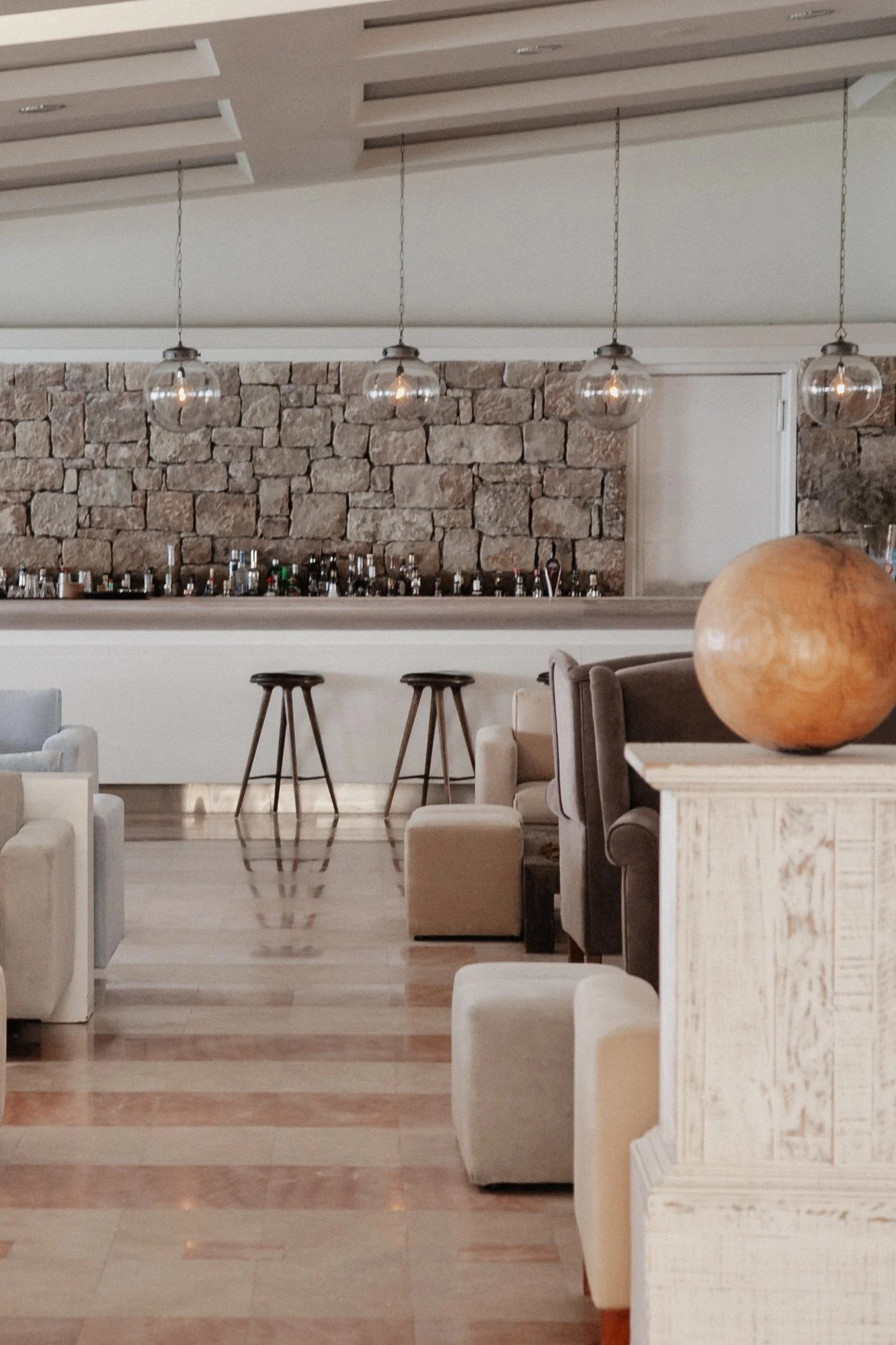 Interior of a lounge area with stone wall, hanging light fixtures, bar counter, stools, sofas, and a large decorative bowl on a white wooden sideboard.