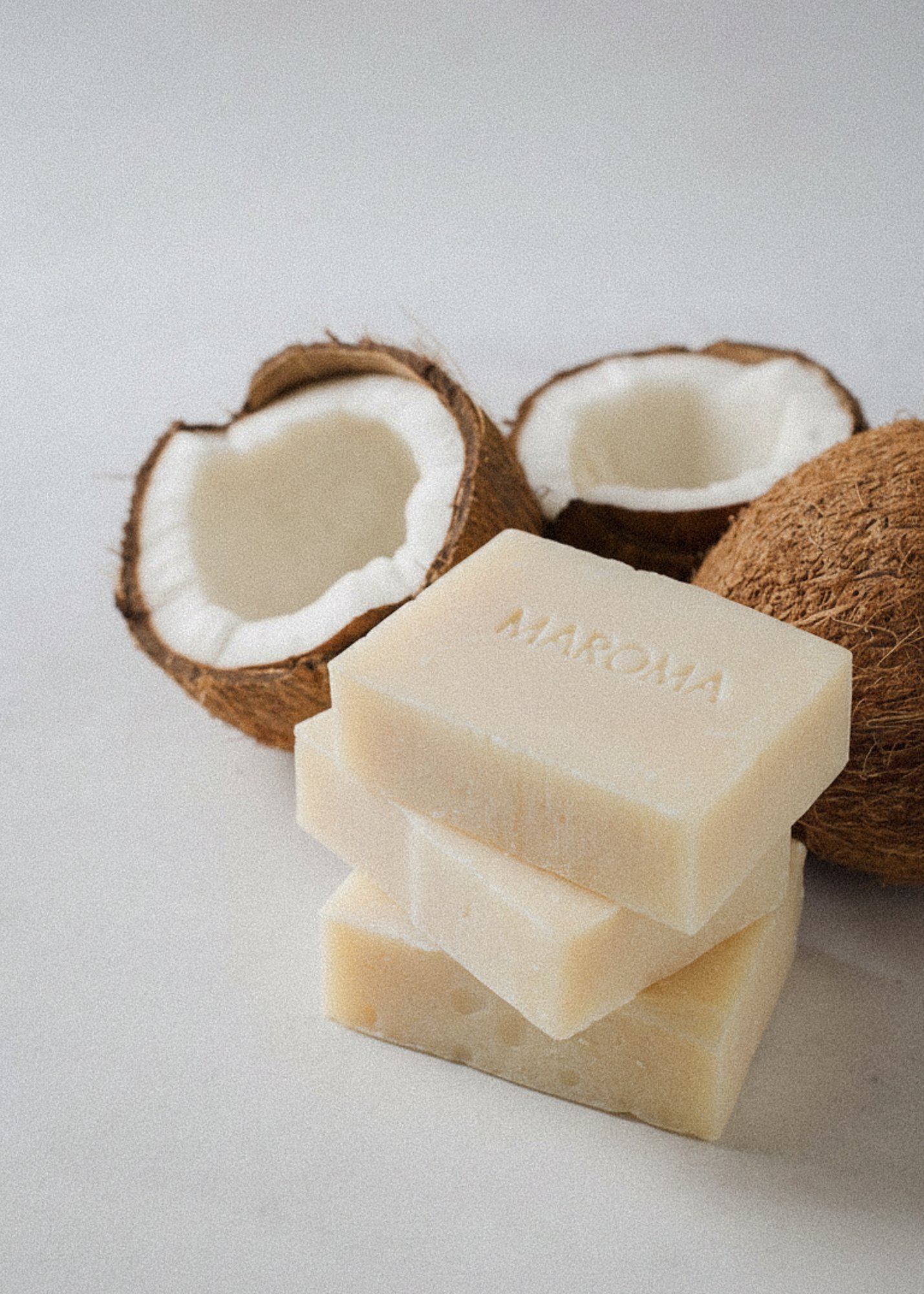 A bar of white soap with the word 'AHAKUYA' inscribed on it, stacked with two additional soap bars, and two coconuts, one of which is cracked open showing white flesh inside.