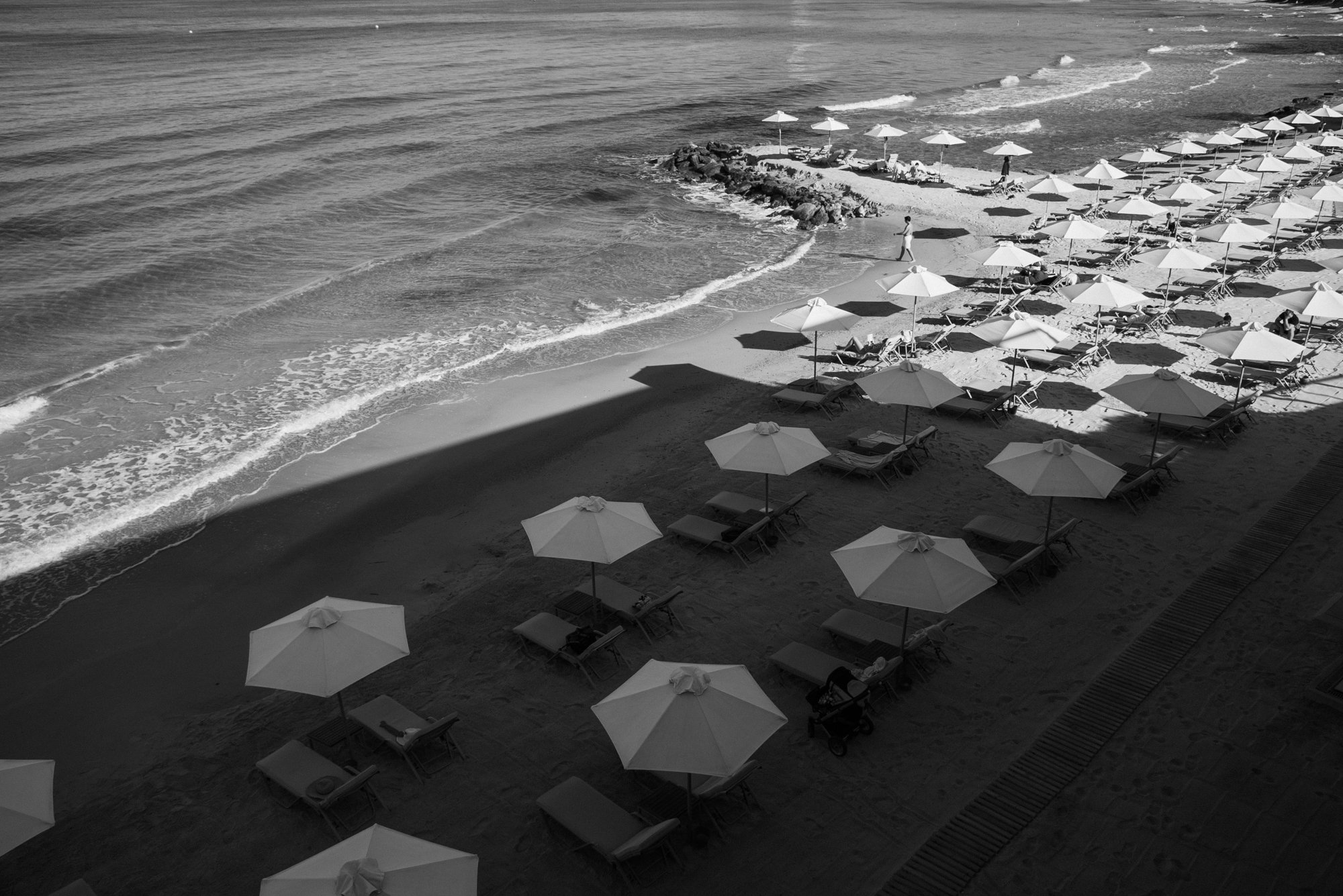 Empty beach with numerous closed umbrellas and lounge chairs, some with towels, along the shoreline with gentle waves, seen in black and white.