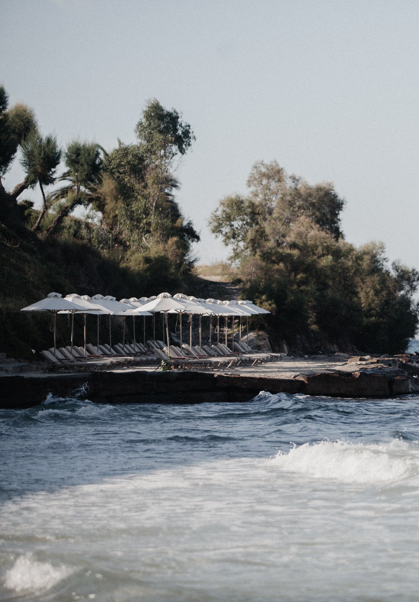 Beach with white umbrellas and lounge chairs along the shoreline, with trees and a hillside in the background.