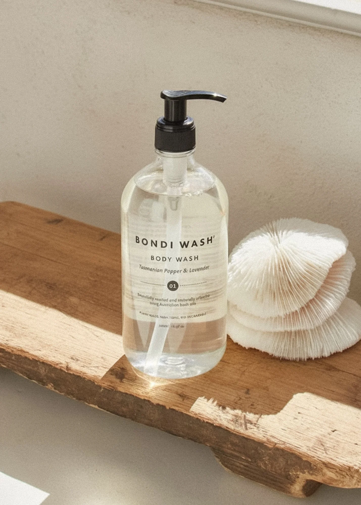 A clear pump bottle labeled "Bondi Wash Body Wash with Australian Pepper & Lavender" on a wooden surface next to a white scrub sponge.