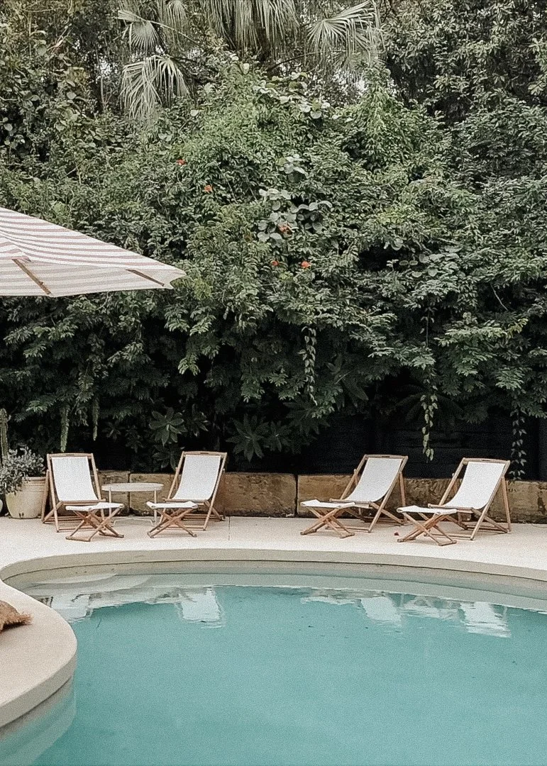 Poolside area with four white lounge chairs with wooden frames, an umbrella, and lush green foliage in the background.