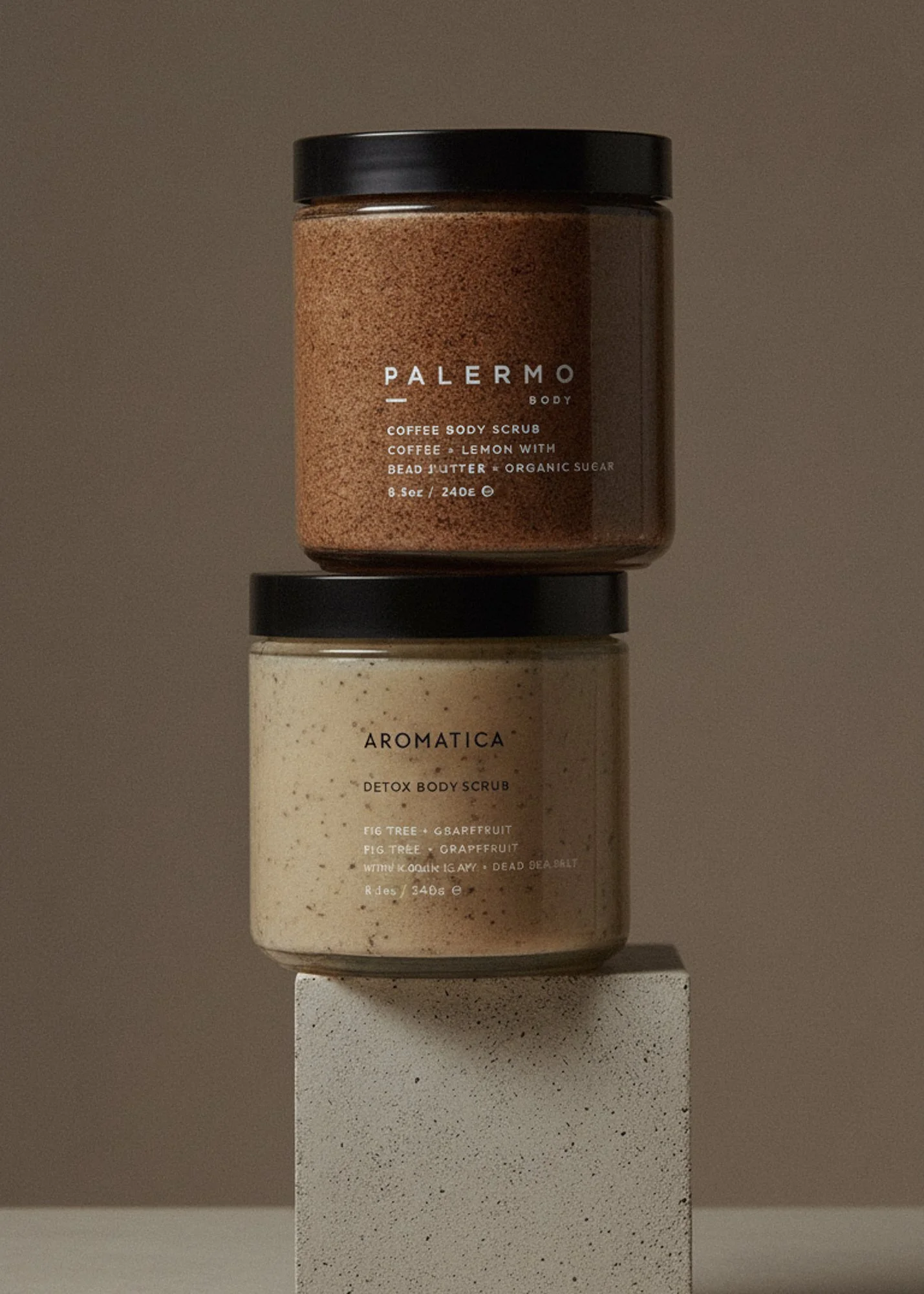 Two jars of body scrub stacked vertically on a white pedestal, one labeled 'Palermo' coffee body scrub and the other 'Aromatica' detox body scrub, against a neutral background.