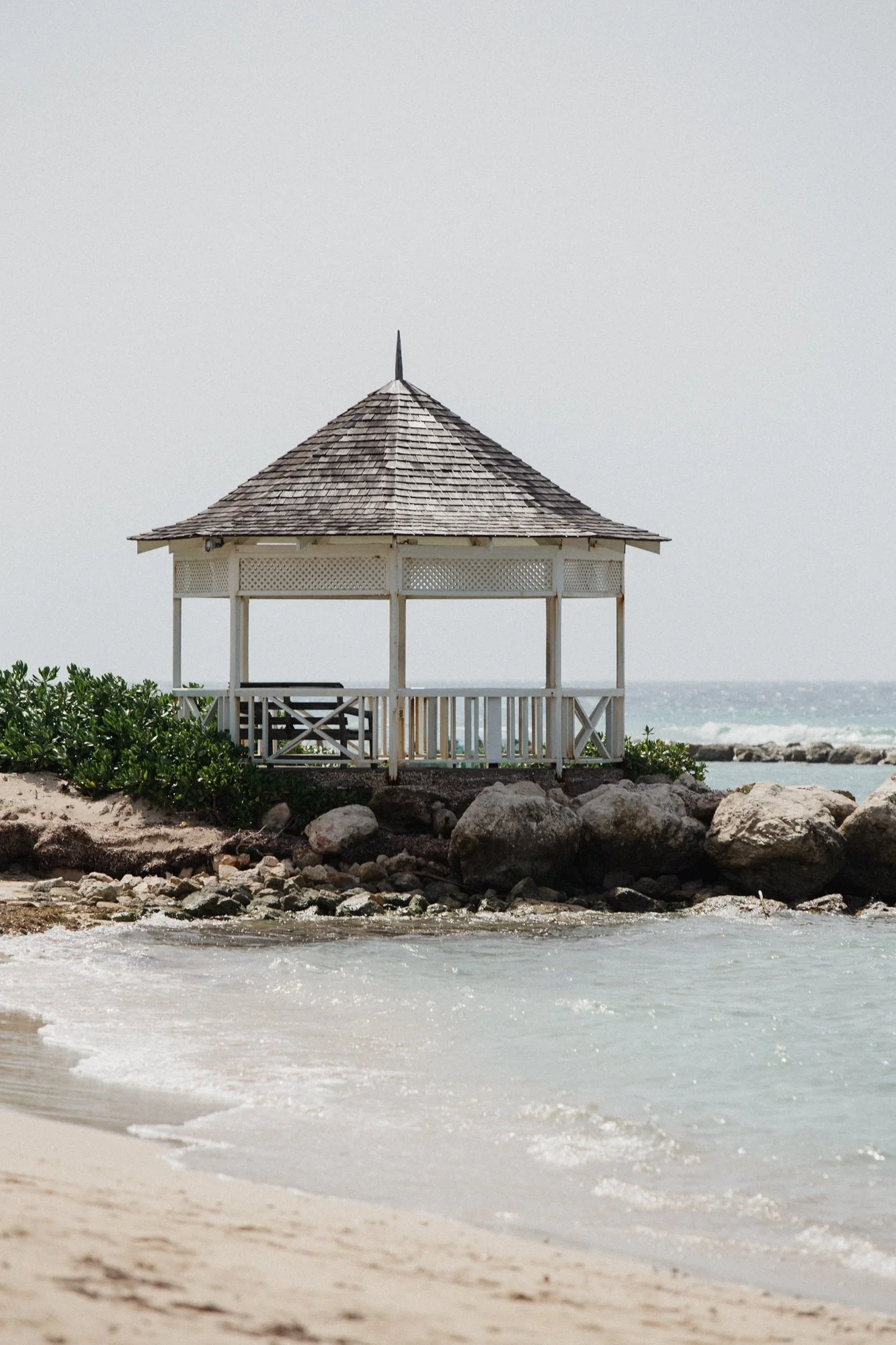 A small white gazebo with a wooden shingle roof on a sandy beach, surrounded by rocks and green foliage, with the ocean in the background.