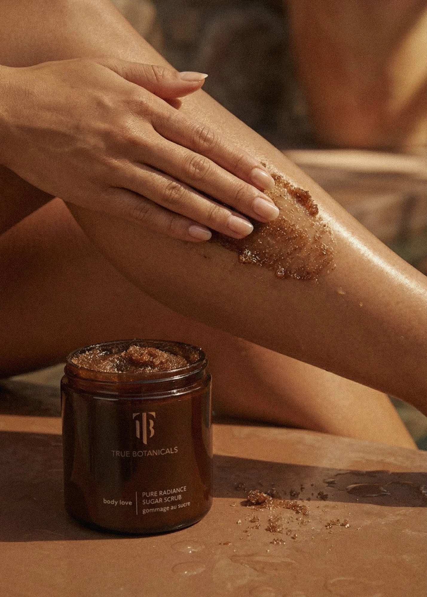 A person applying sugar scrub to their leg. The scrub is from True Botanicals and labeled 'Pure Radiance Sugar Scrub'. The person is using their hand to massage the scrub onto their skin, with some of it spilled on a wooden surface.