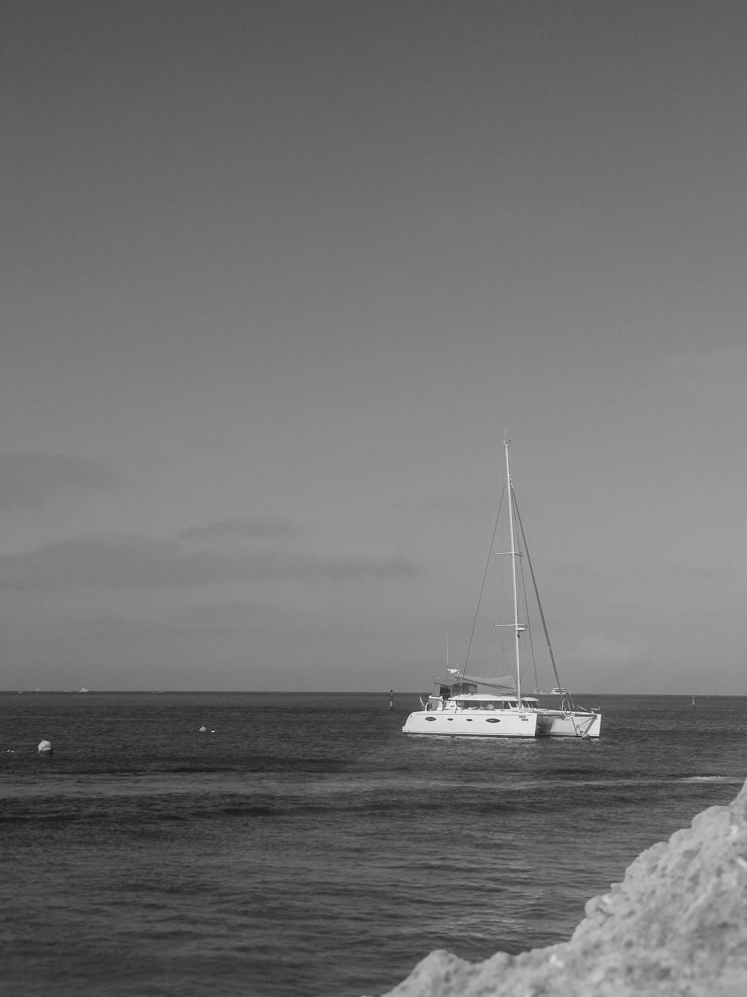 A sailboat anchored in calm water near a rocky shore on a clear day with a partly cloudy sky.