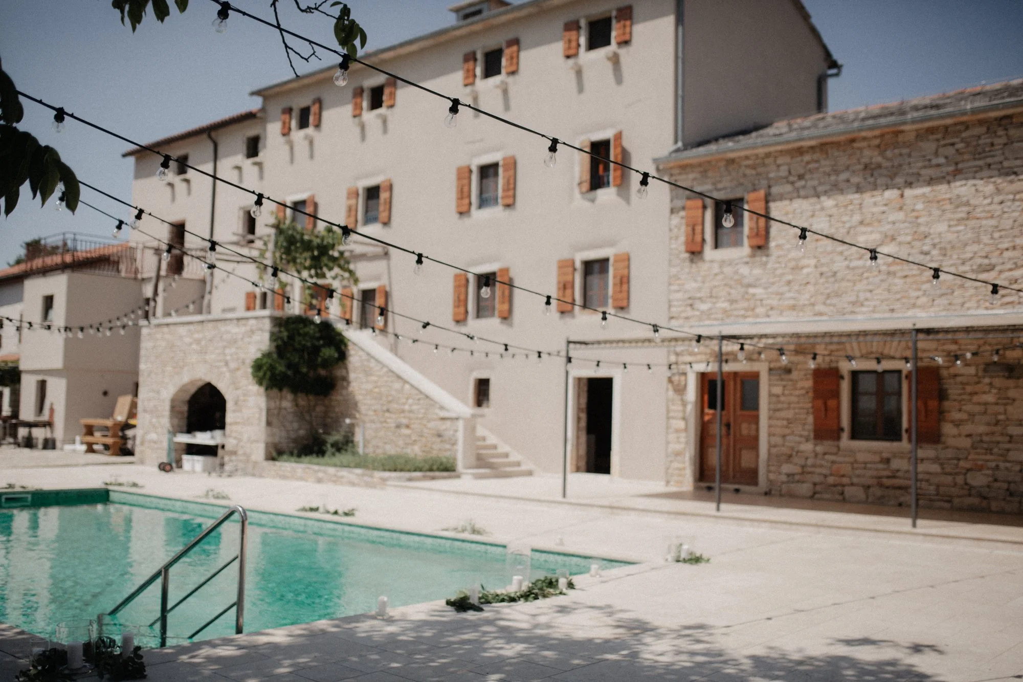 An outdoor pool area in front of a villa-style building with stone and stucco walls, wooden shutters, and string lights hanging overhead.
