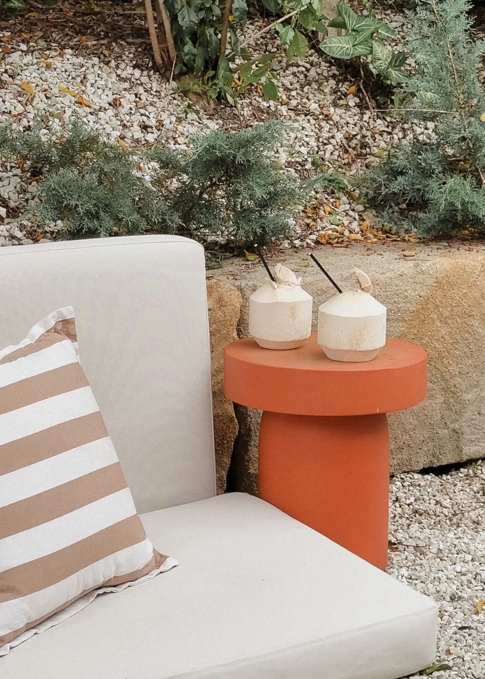 Outdoor scene with a white cushioned seat, a striped beige and white pillow, a terra-cotta colored side table, and two coconut drinks with straws on the table. In the background, there are green plants and rocks.