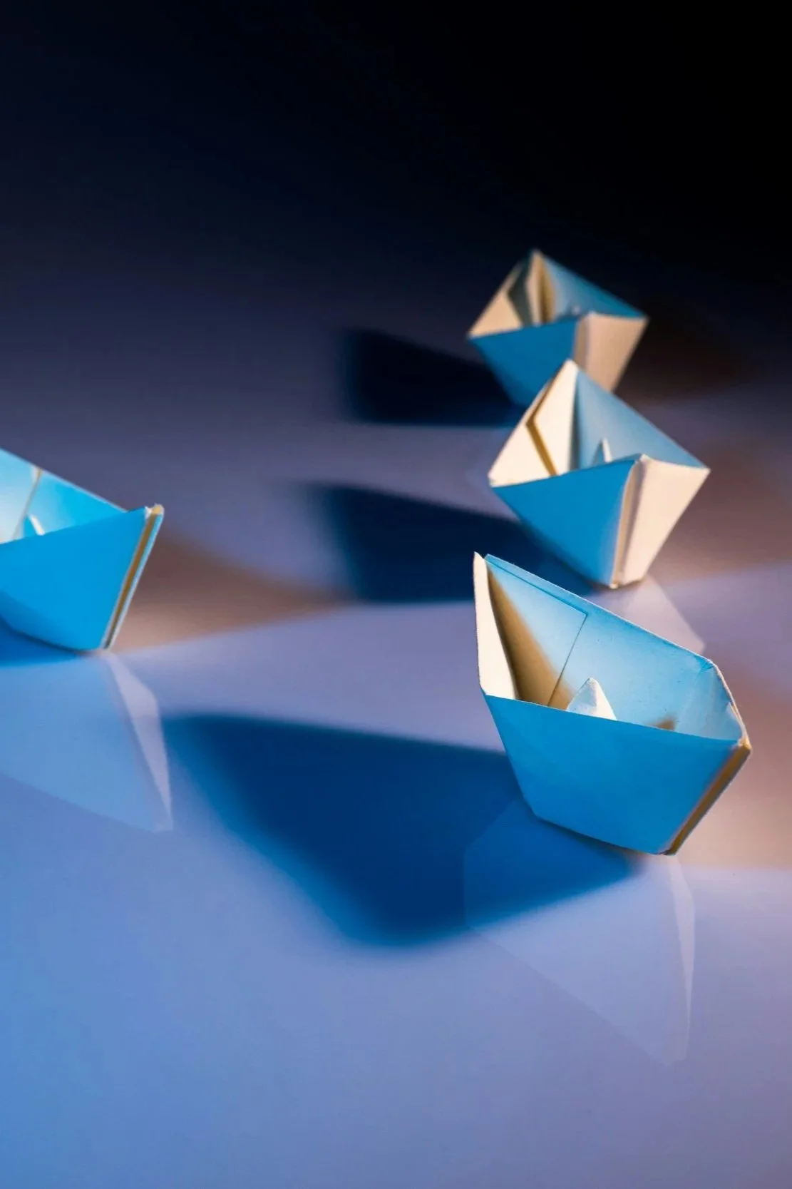 Fünf Papierschiffe in blauer und weißer Gestaltung auf einer glatten Oberfläche, mit Licht- und Schattenspielen.