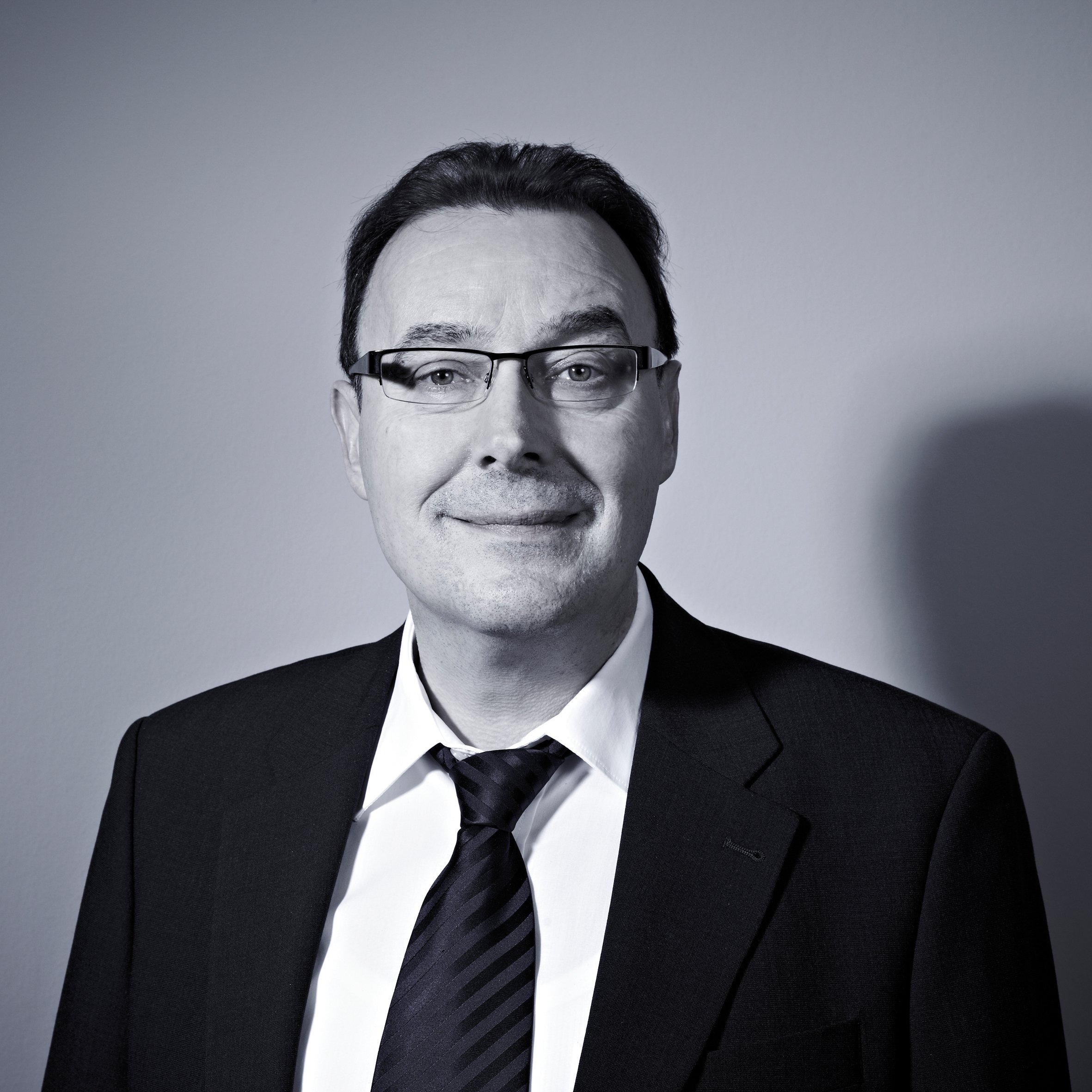 Schwarz-weiß-Fotografie eines Mannes in Anzug, Krawatte und Brille, lächelnd, vor neutralem Hintergrund