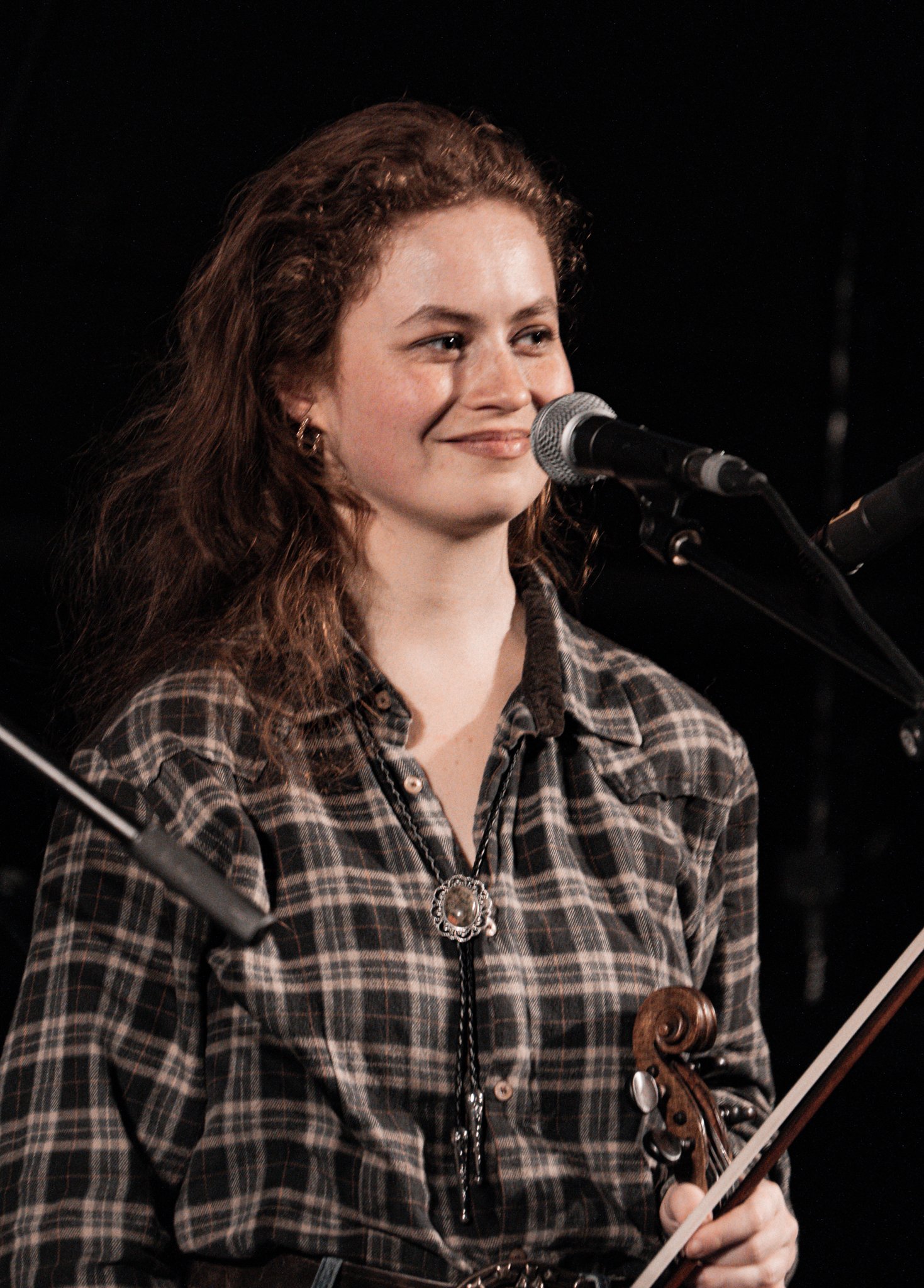 A woman with curly red hair sits in front of a microphone, holding a violin, wearing a plaid shirt and a decorative necklace, smiling softly.