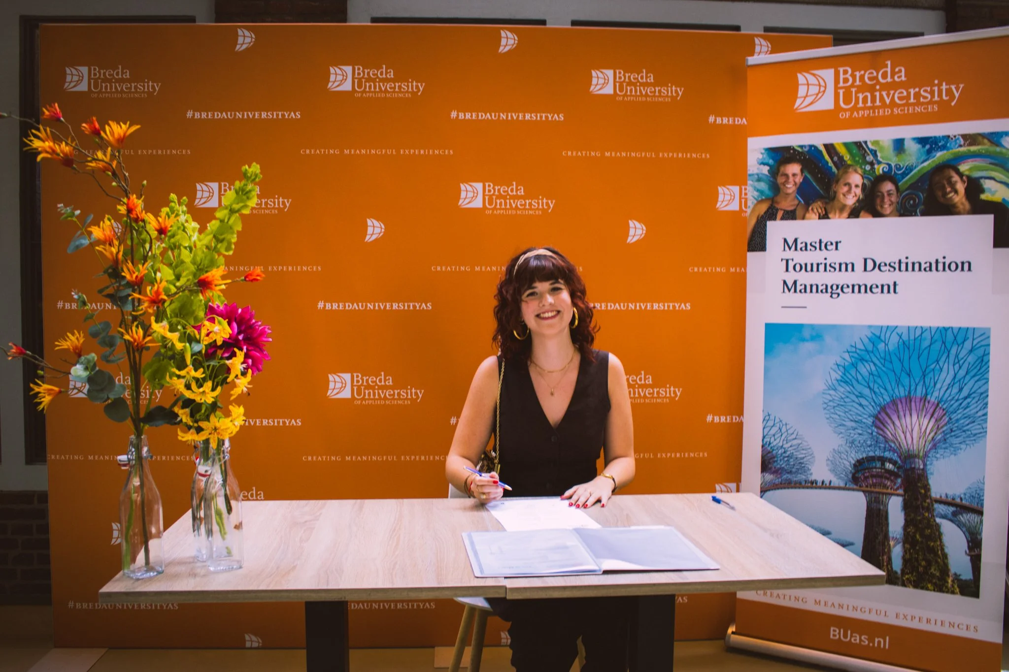 A woman sitting at a table with an open folder, holding a pen, and smiling. Behind her, an orange backdrop with the Breda University logo and hashtags. To her left, a bouquet of colorful flowers in glass vases. To her right, a poster advertising a Ma