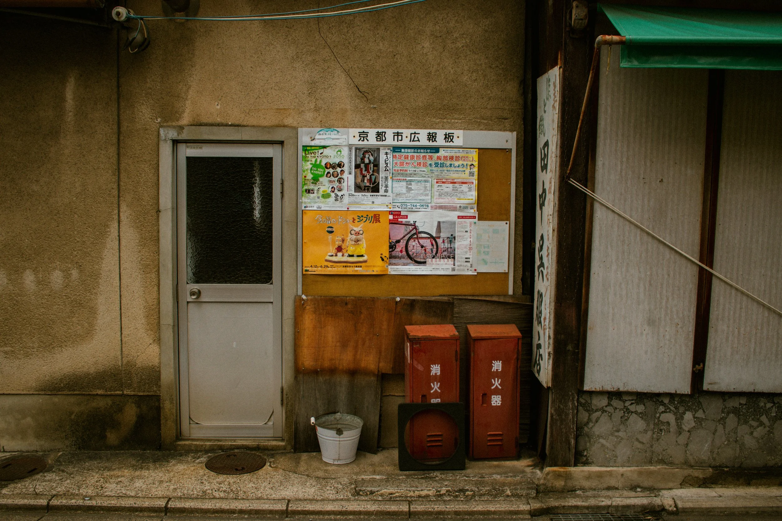 A wall with a door, bulletin board with posters, two red fire extinguishers, a small white bucket, and a black box on the sidewalk in front.