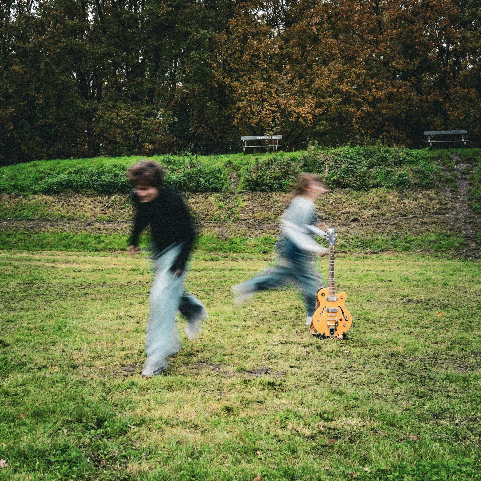 Two children playing outdoors near a guitar standing on the grass, with trees and benches in the background, blurred as they run.