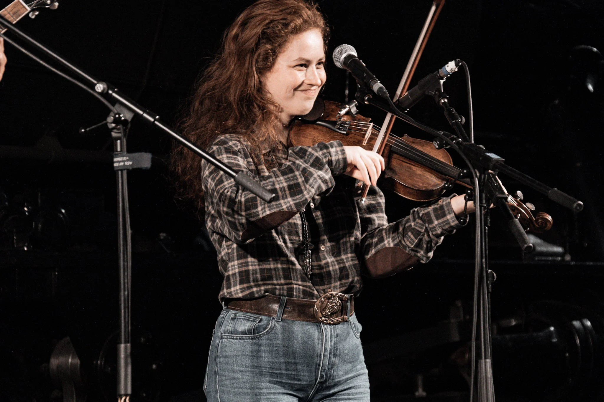 A woman with long, curly hair playing the violin on stage, wearing a plaid shirt and jeans, with multiple microphones set up around her.