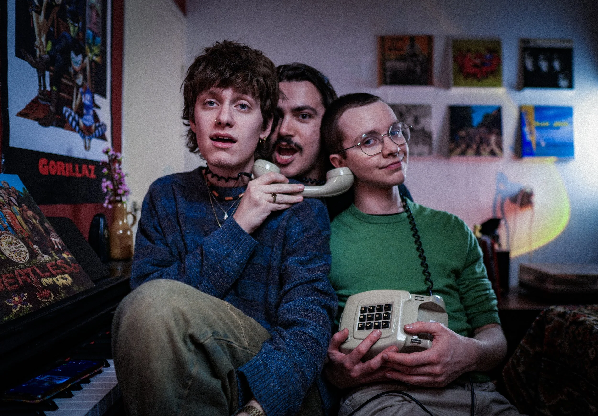 Three young men in a room, one holding a vintage phone, another with a phone on his neck, and the third with a phone in their lap, surrounded by music posters and artwork.
