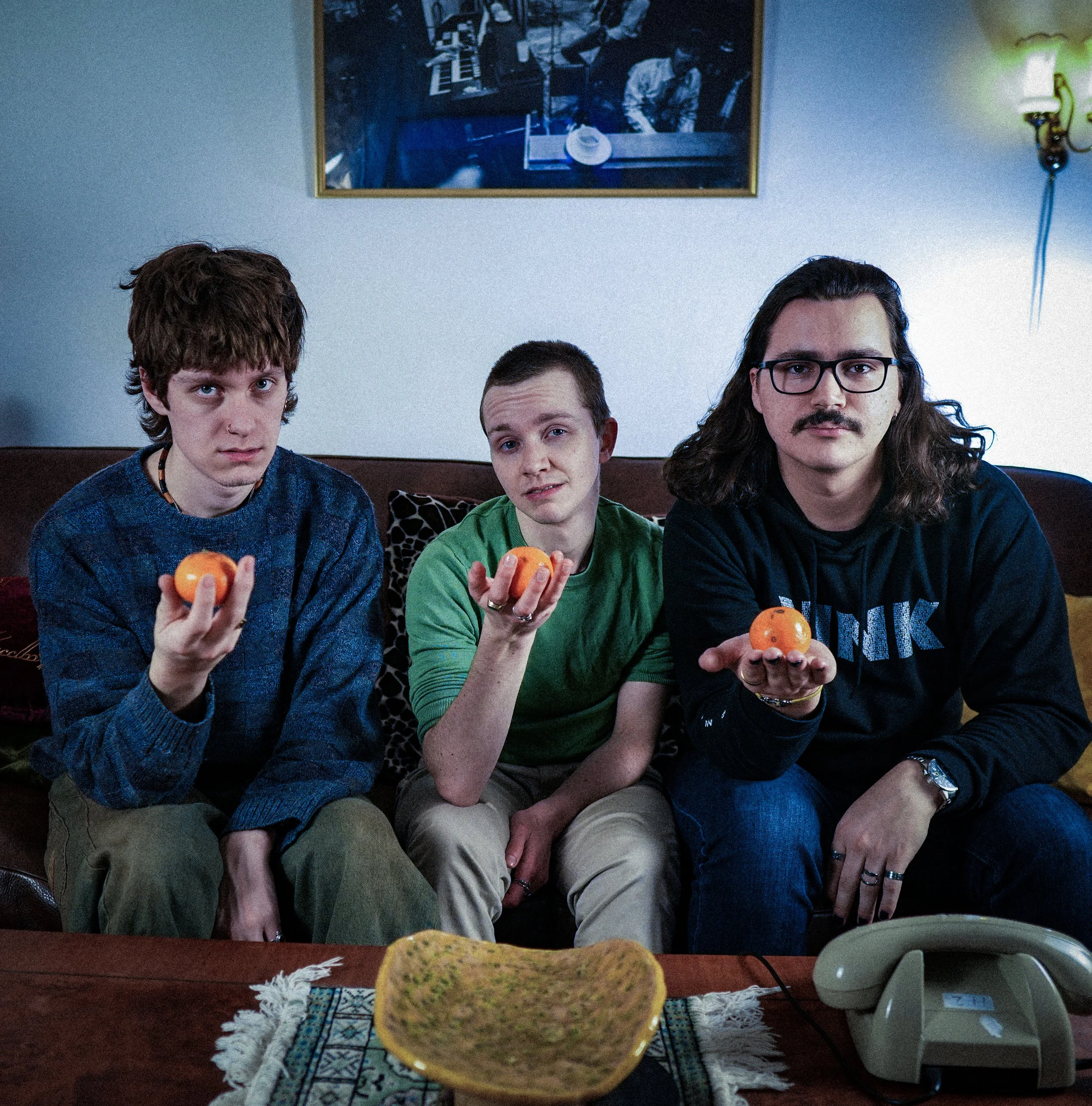 Three young men sitting on a couch, each holding an orange in their hand, with a bowl on the table in front of them, and a vintage phone.