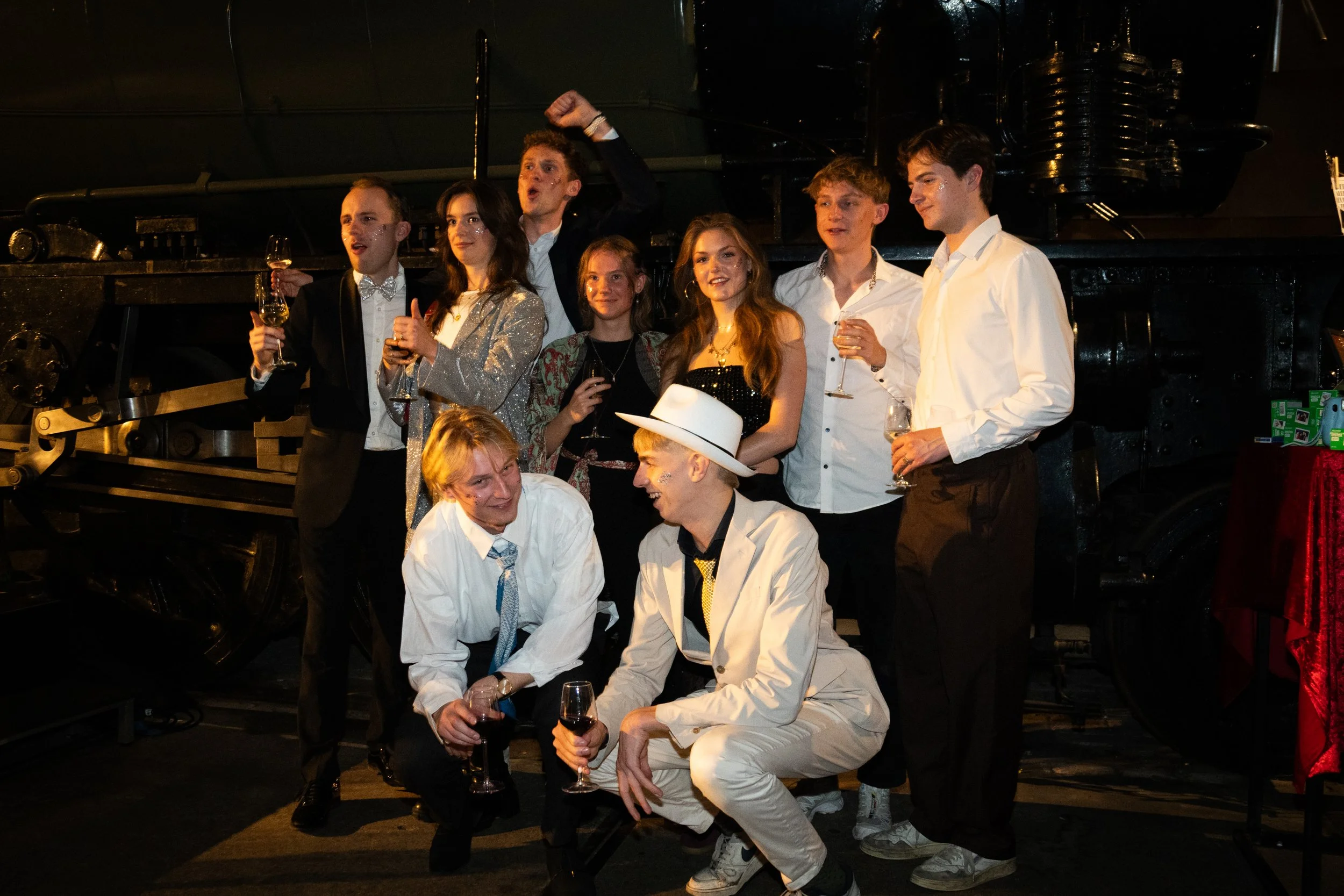 A group of people dressed in formal and semi-formal attire, celebrating with drinks in front of a large black vintage steam locomotive.