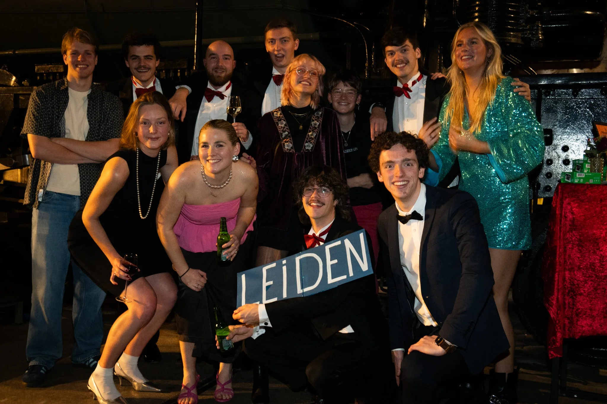A group of young adults and a woman with blonde hair, dressed in formal attire, posing together at a party or celebration. They are smiling and holding drinks, with one person holding a sign that says 'Leiden'.