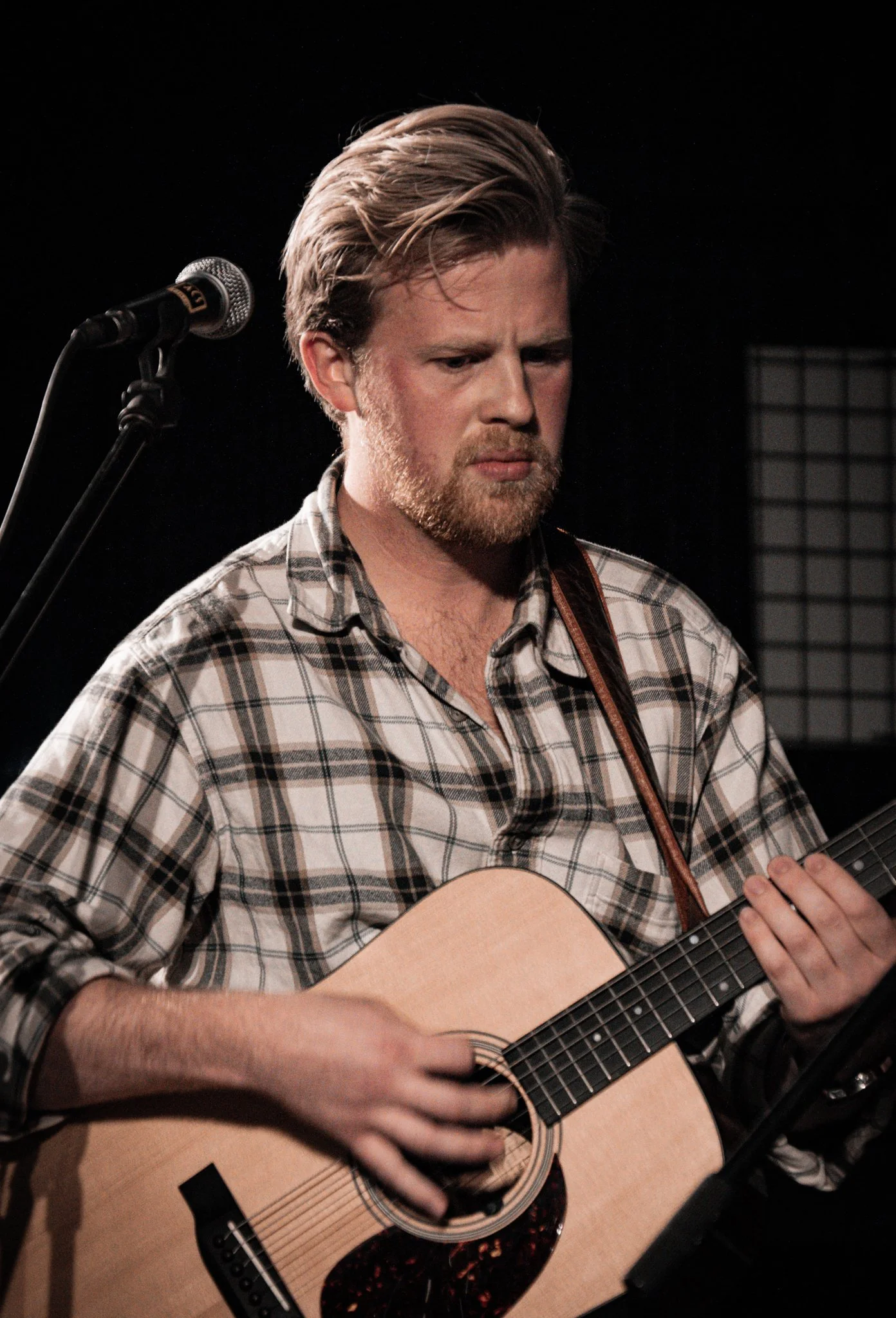 A man with blonde hair and a beard, wearing a plaid shirt, playing an acoustic guitar on stage.