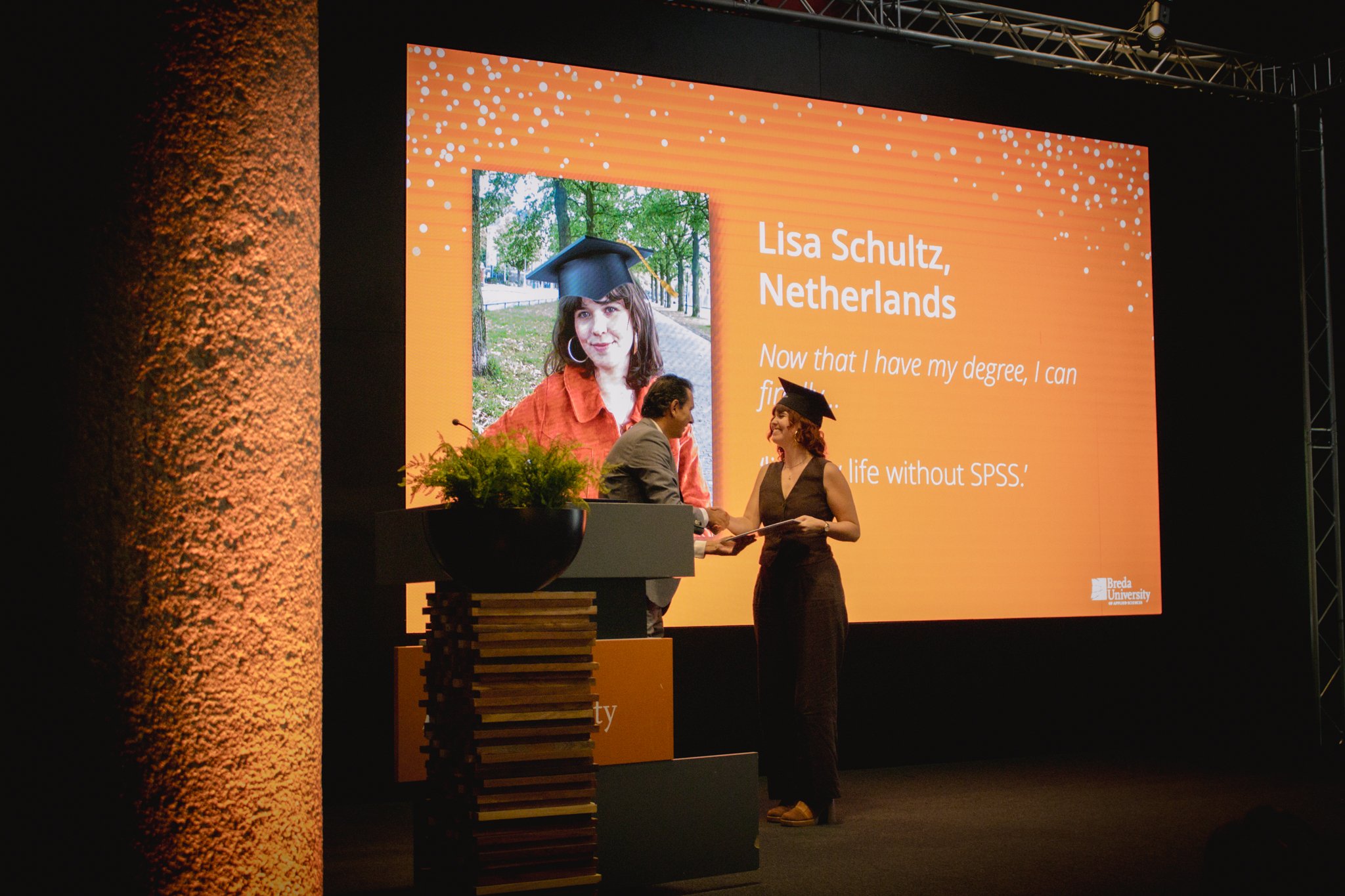 A woman in a black dress and cap receives a diploma on stage from a man in a gray suit. They are shaking hands, with a large orange background screen behind them displaying a photo of the woman and information about her. The screen mentions her name,