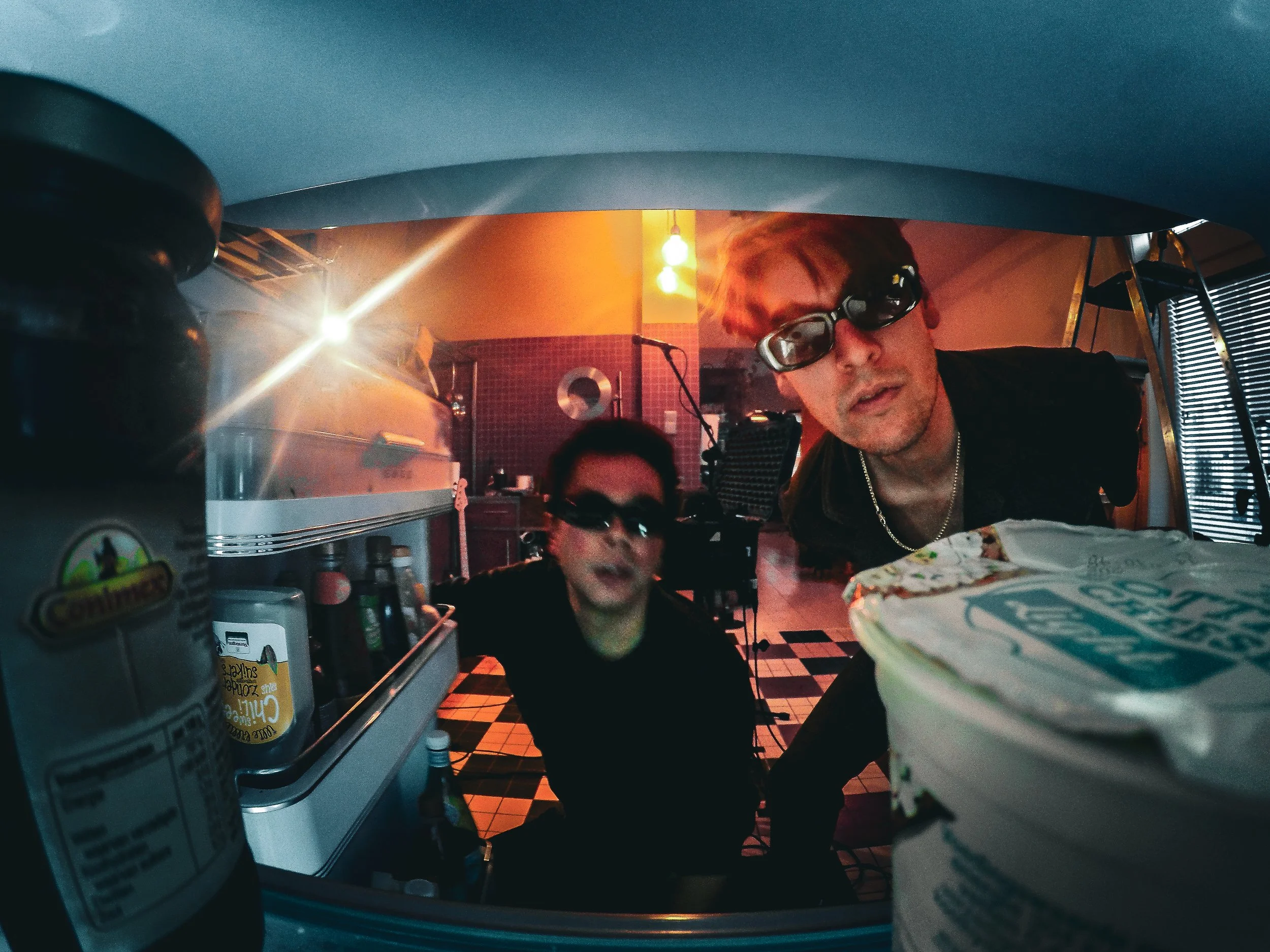 Two men looking into a refrigerator, wearing sunglasses, with various food items and condiments inside, in a retro-style kitchen with checkered floor and warm lighting.