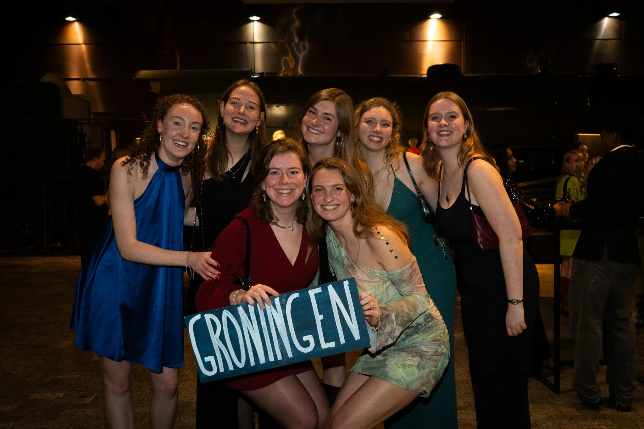Group of eight young women in formal attire posing together at an indoor event, holding a blue sign that says "Groningen".