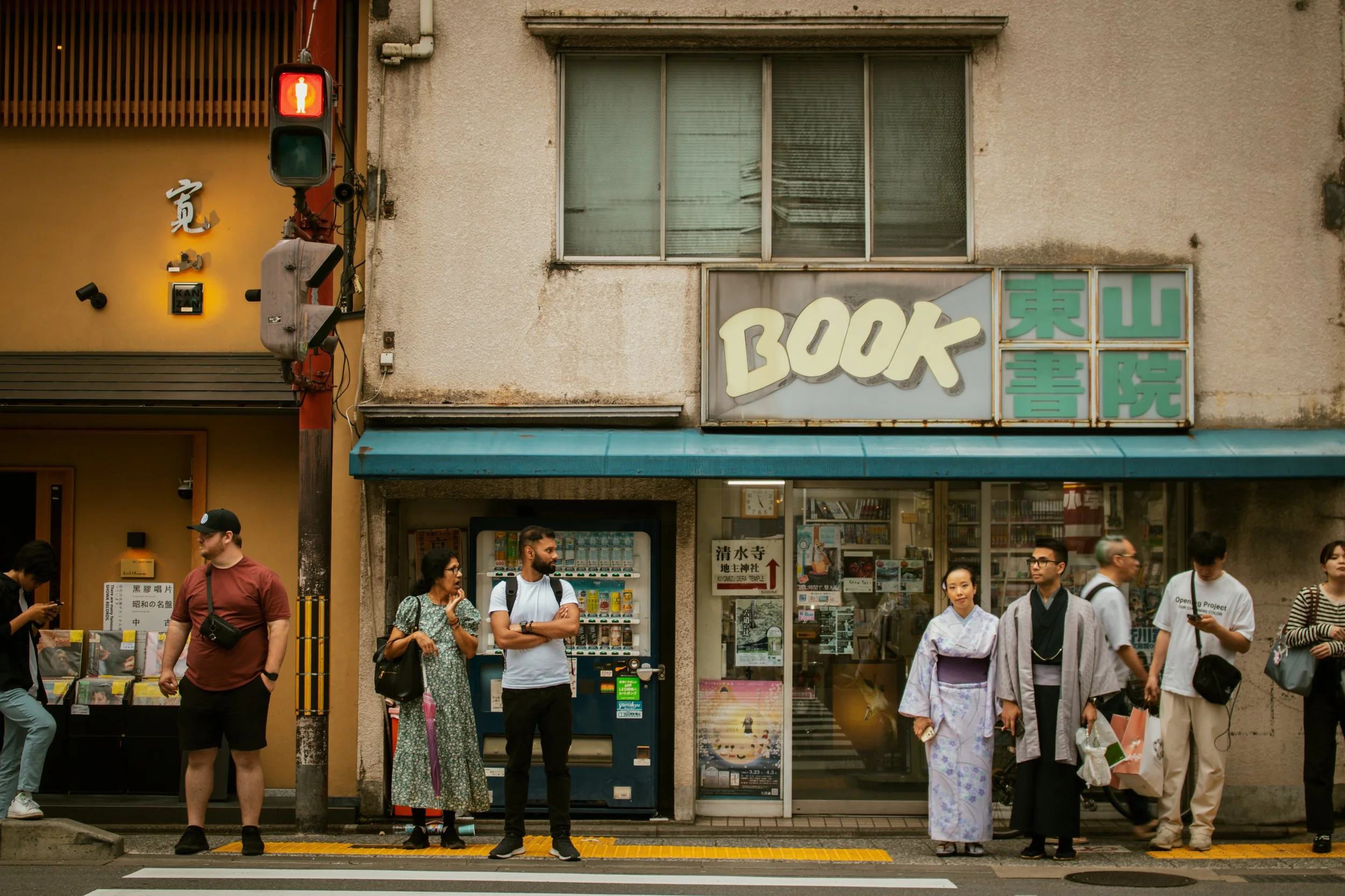 People waiting at a crosswalk in front of a bookstore with a sign that reads 'BOOK' and Japanese characters, a red traffic light, and a vending machine outside.