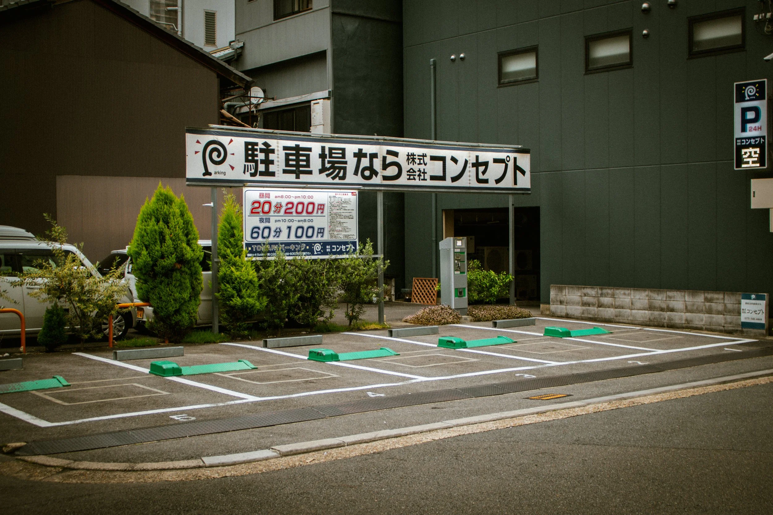 Empty parking lot with designated parking spaces, a parking payment kiosk, and signs in Japanese indicating parking rates and hours, surrounded by greenery and nearby buildings.