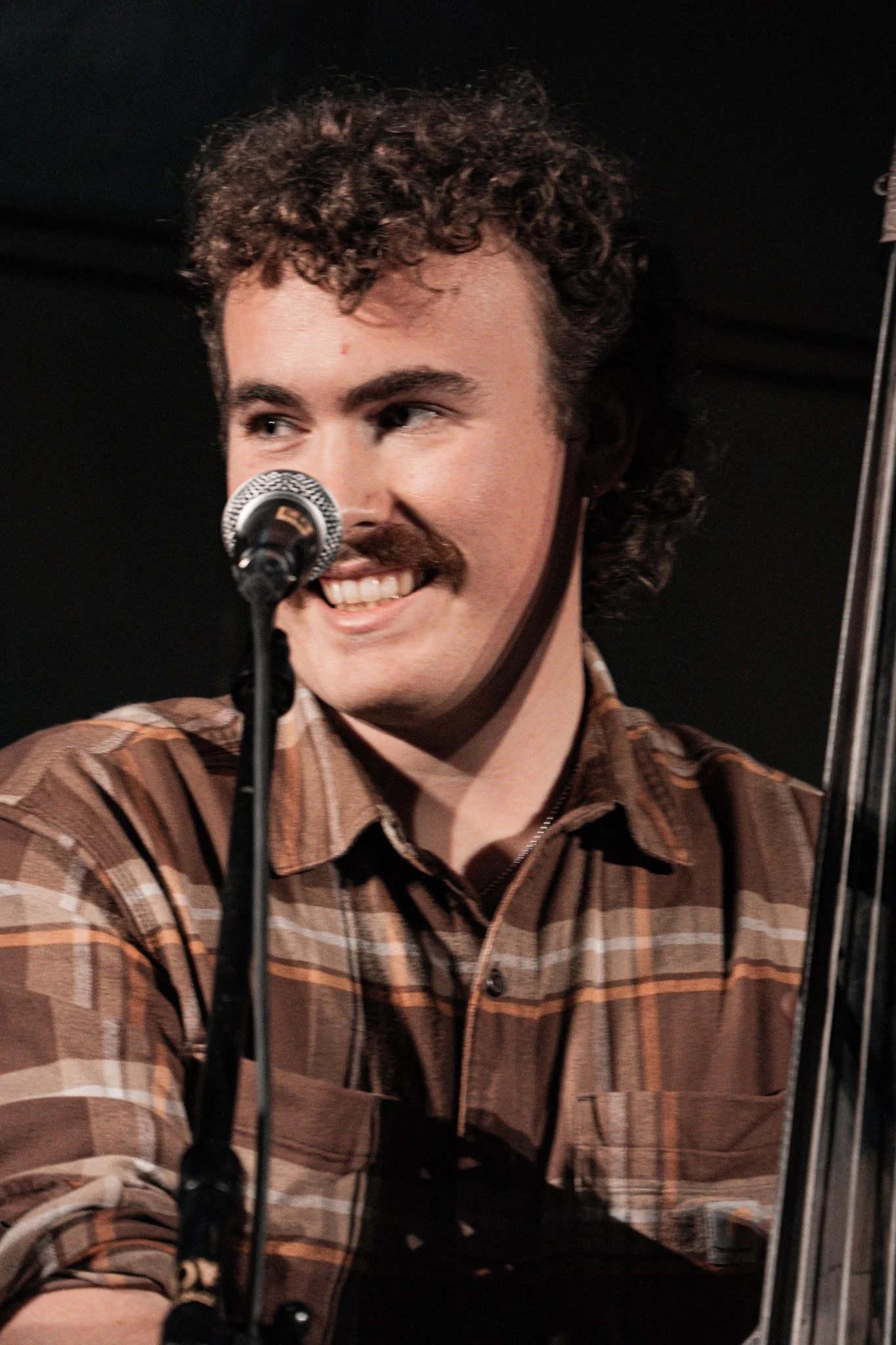 A man with curly hair and a mustache speaking into a microphone, wearing a brown plaid shirt.