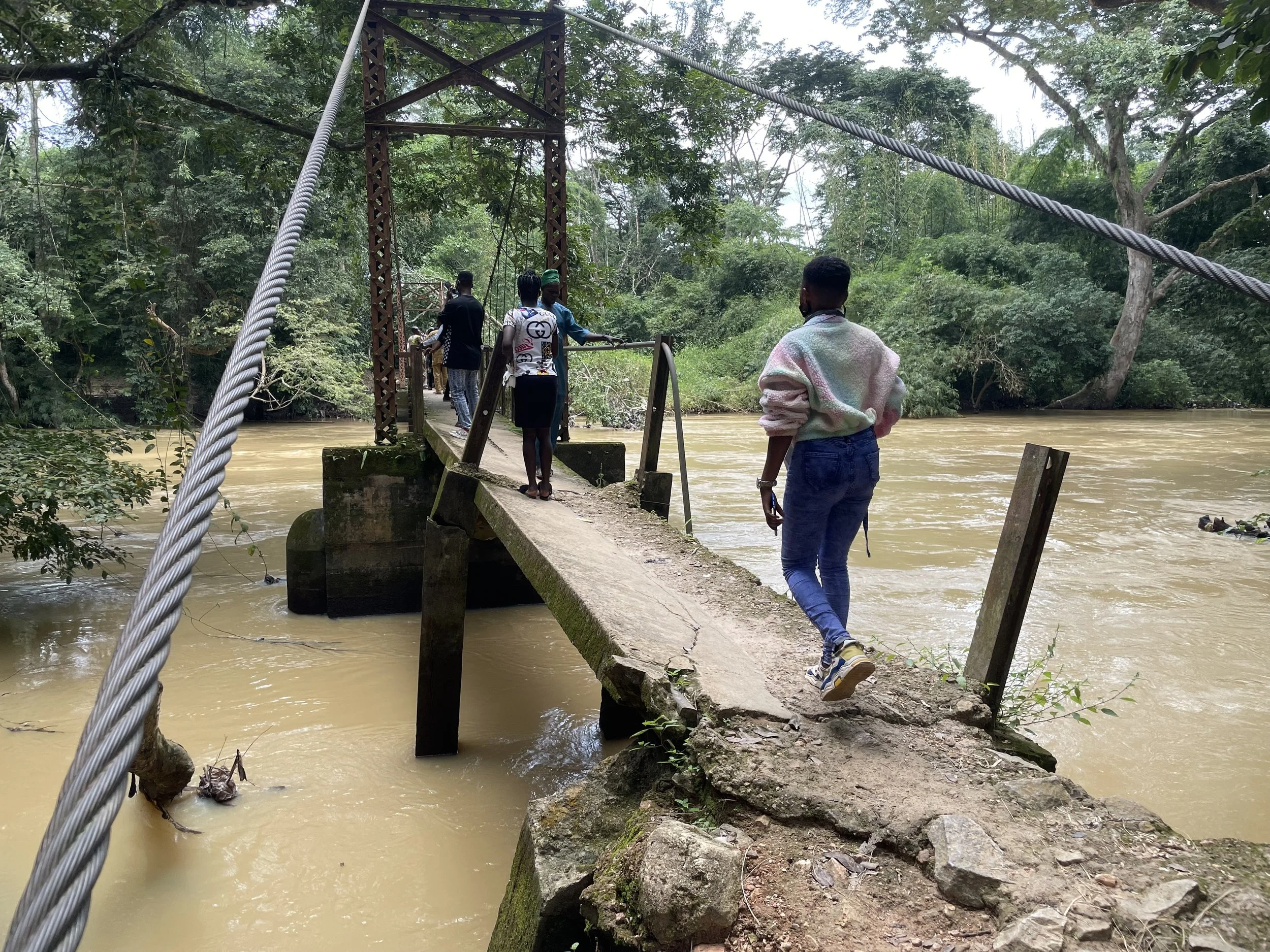 People crossing a small suspension bridge over a muddy river surrounded by green trees.