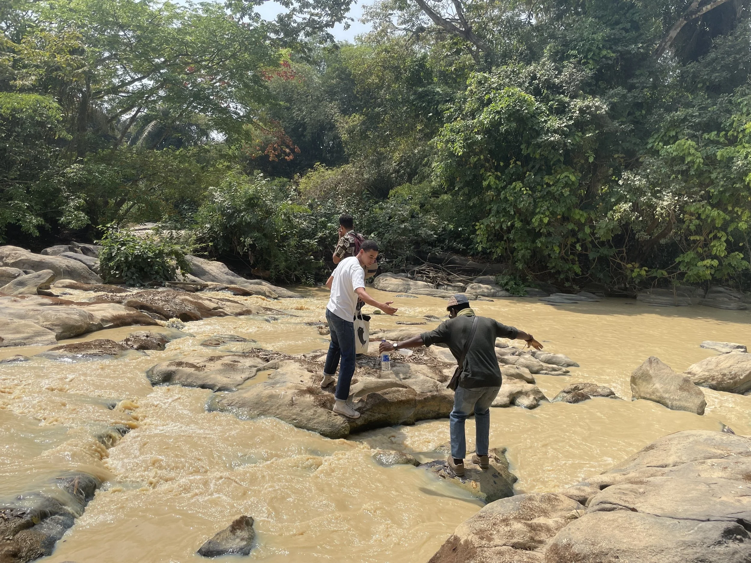 Three people crossing a rocky stream in a lush, green forest, with one person helping another step onto a rock.