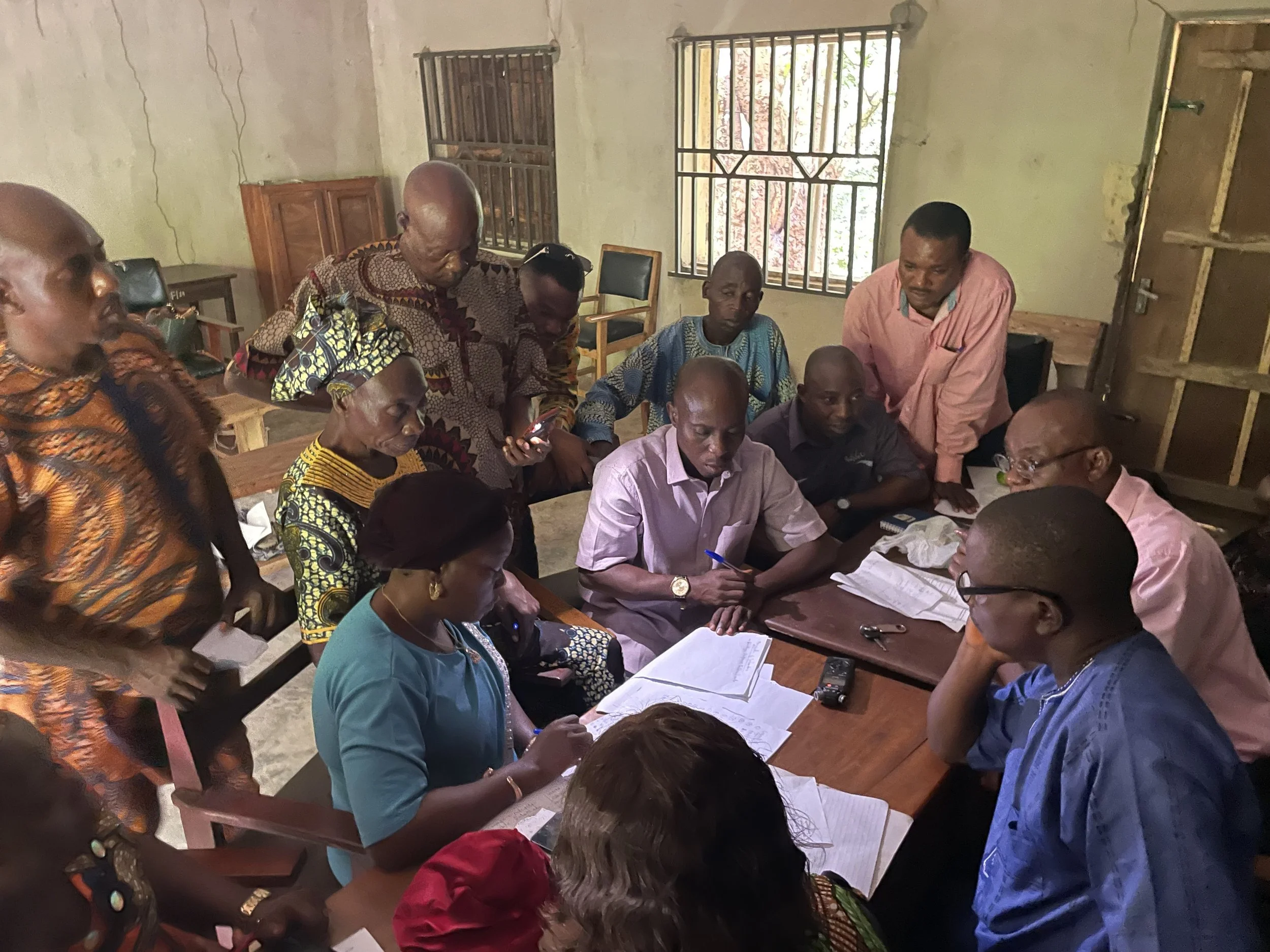 A group of people gathered around a table in a room, engaged in a discussion with documents and a recording device on the table.