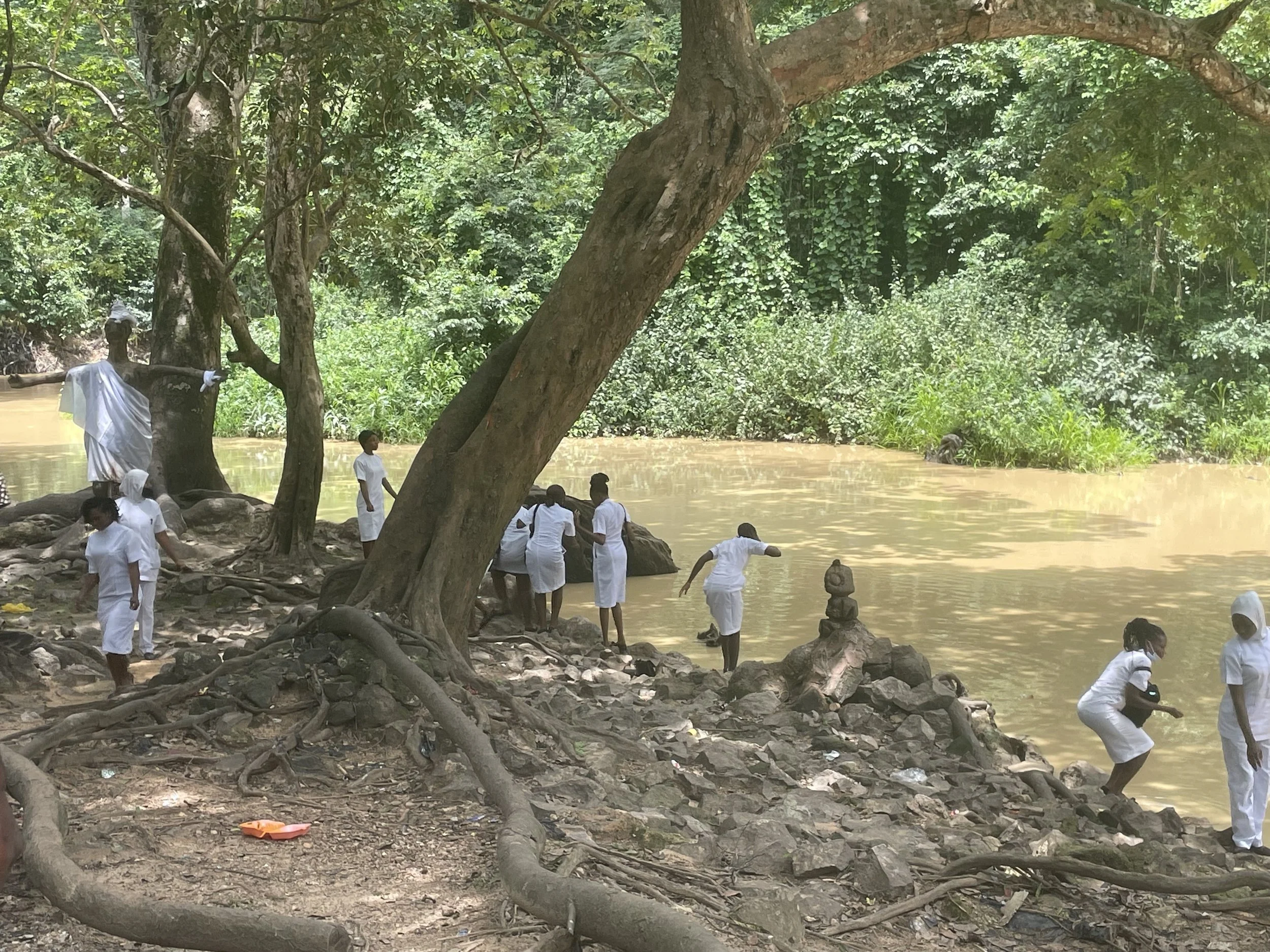 People wearing white clothing gather along a riverbank surrounded by trees, with some   near the water and some sitting or standing on rocks.
