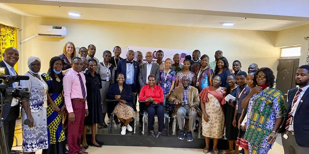 Group photo of diverse professional people in a conference room, some standing and some seated, with banners and a camera in the room.