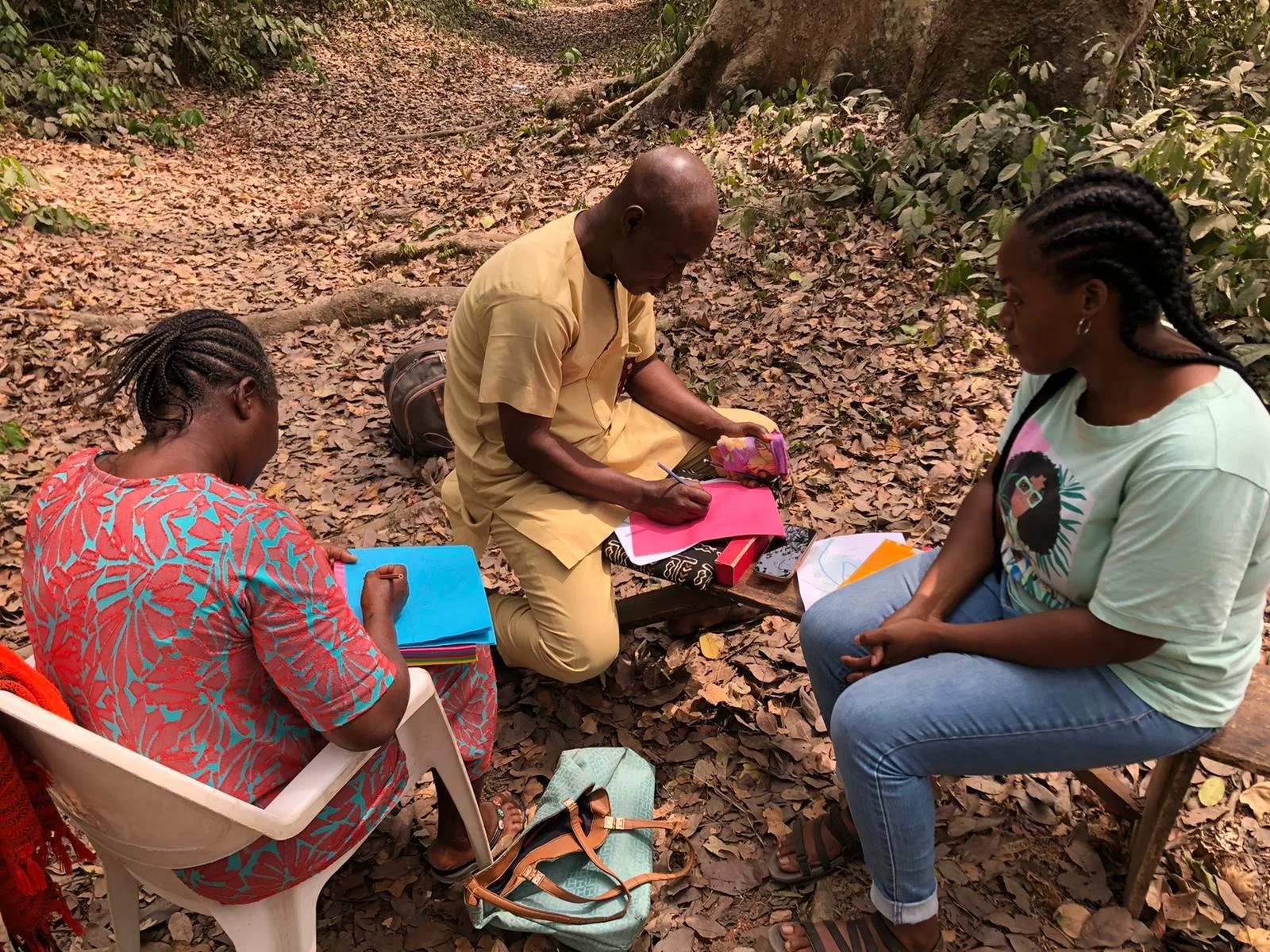 Two women and one man sitting outdoors in a wooded area, engaged in writing or drawing on colorful papers placed on their laps and the ground, surrounded by fallen leaves and green foliage.