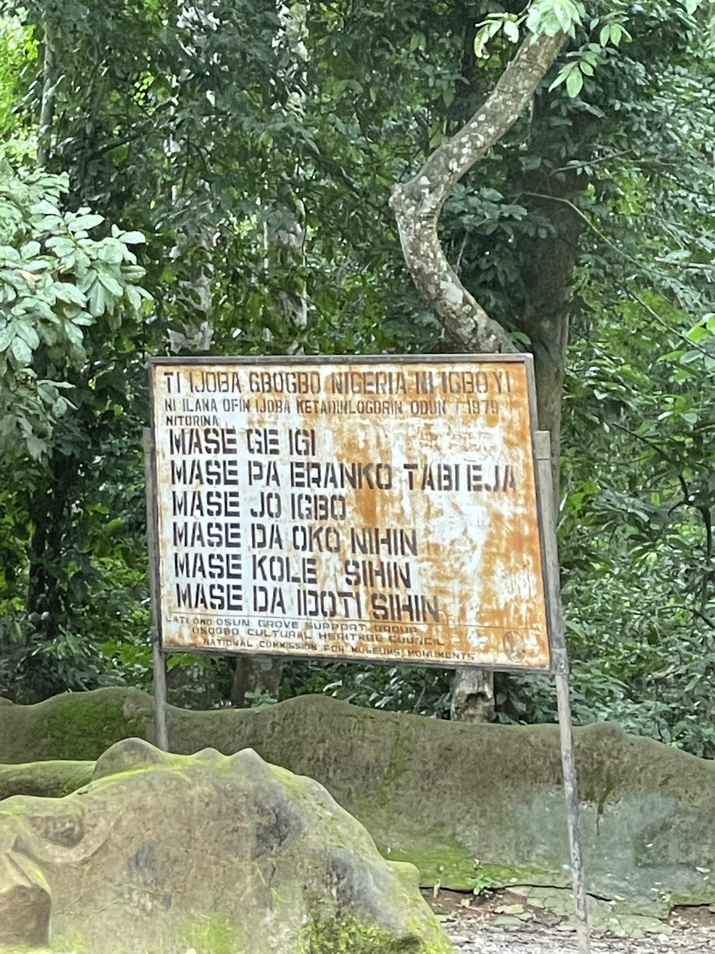 A weathered signboard in a wooded area with trees and rocks in the background, containing text in Yoruba about rules at the Osun-Osogbo Sacred Grove.