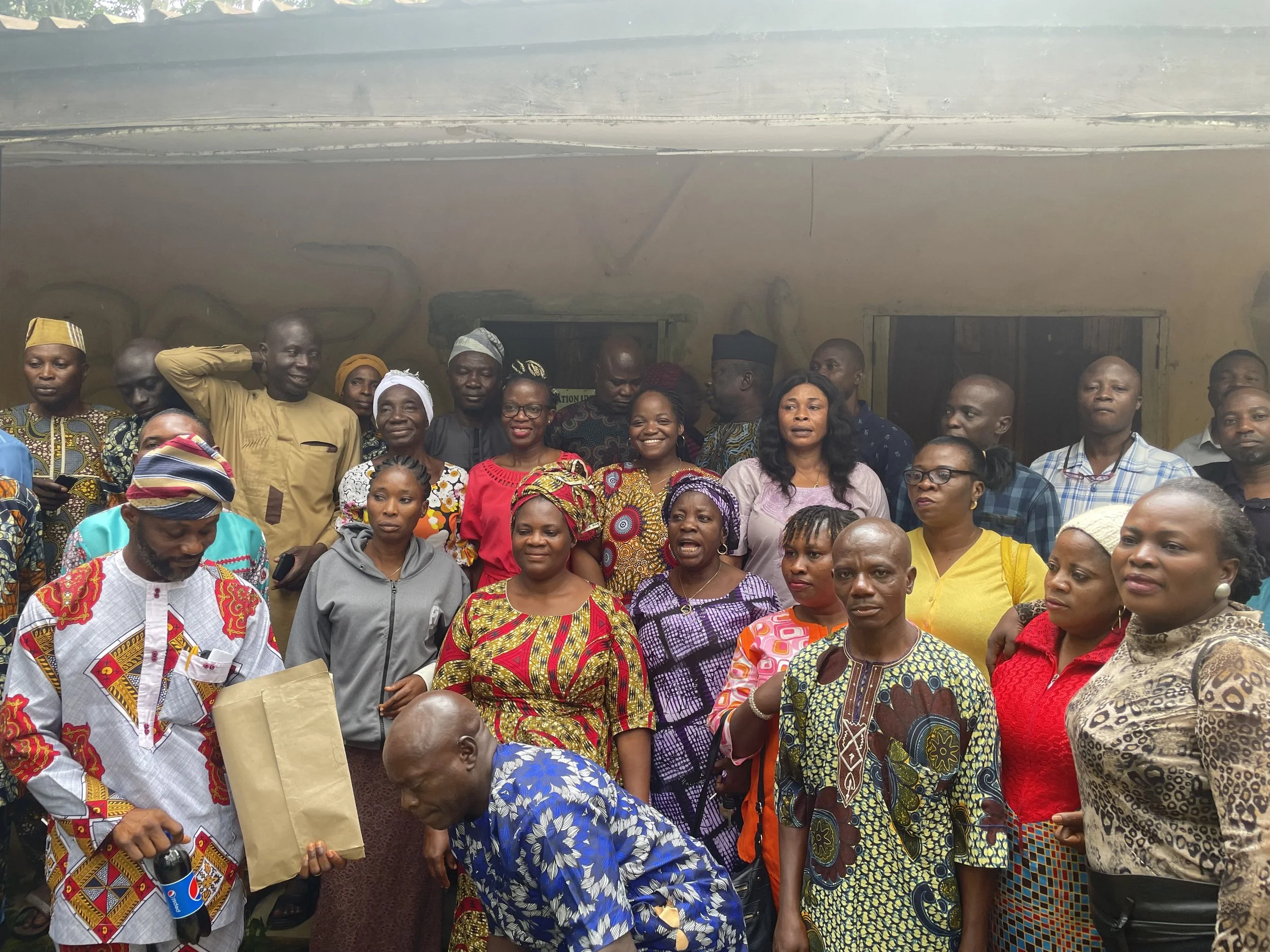A large group of people gathered outdoors, some are smiling and others are listening attentively. The group includes men and women of various ages dressed in colorful traditional and modern clothing.