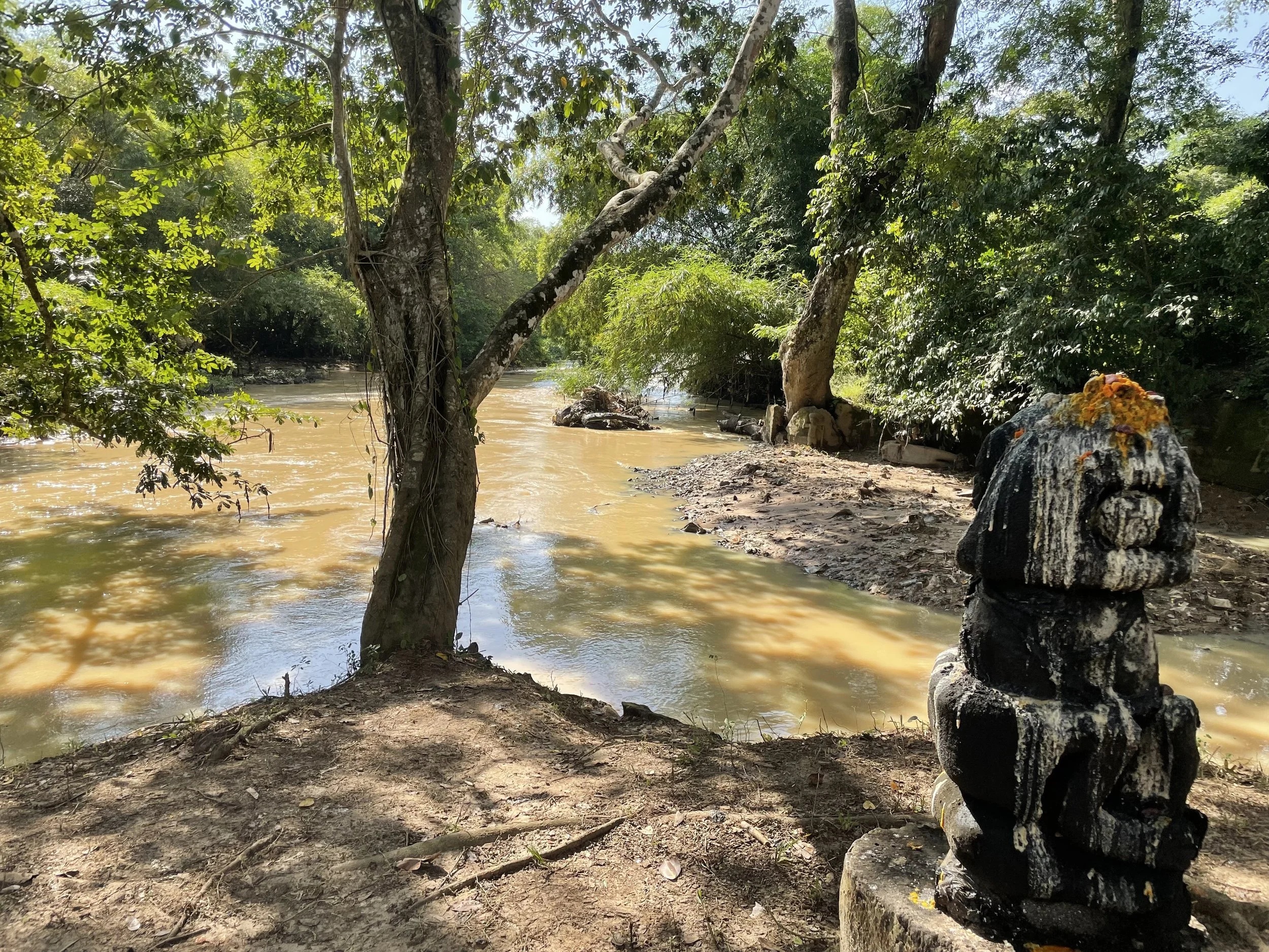 A river flowing through a lush green forest with trees on both sides. A small carved stone statue decorated with libations is in the foreground on the right.