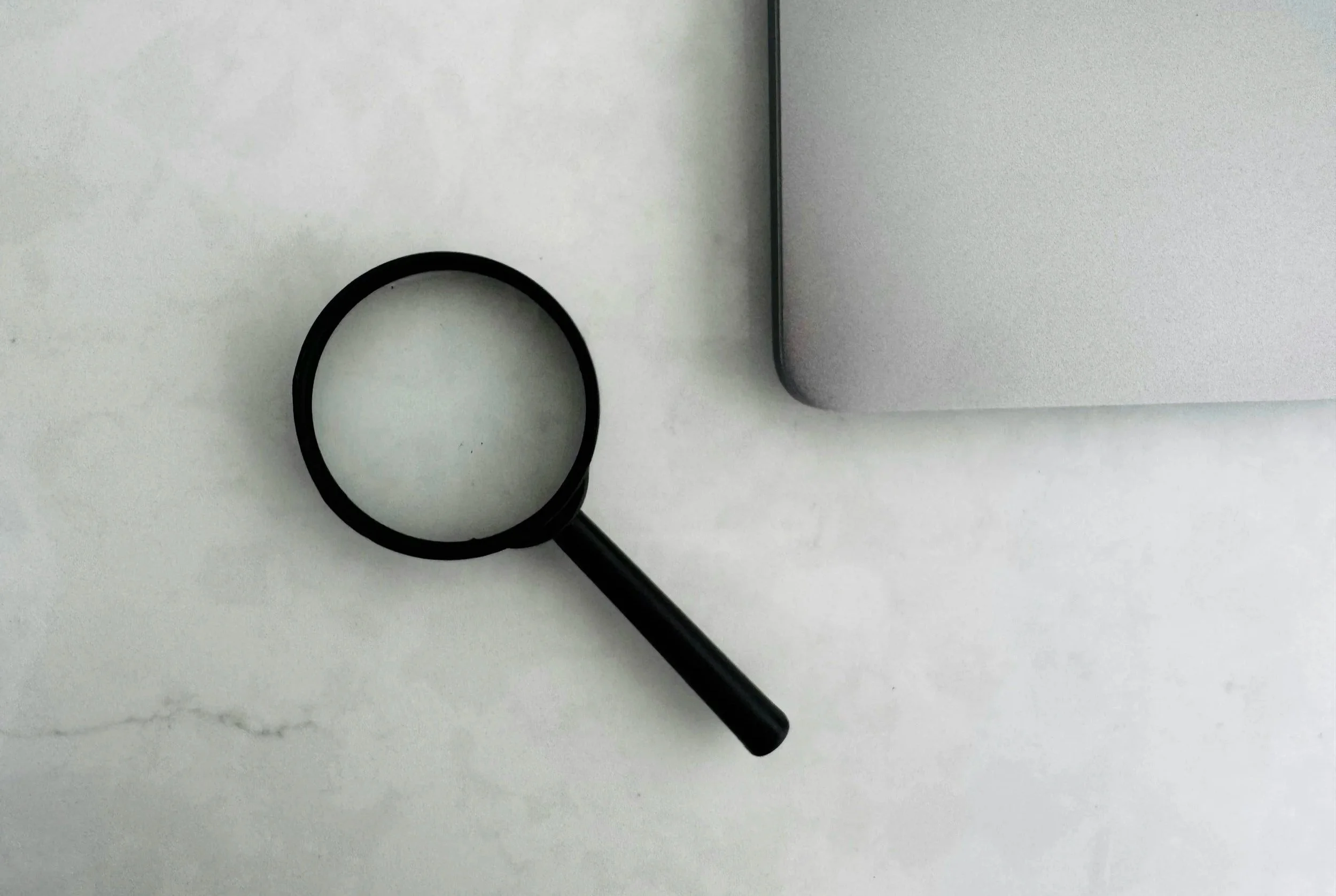 A flat lay photograph of a black magnifying glass, a black circular rubber band, a silver laptop, and a marble surface.