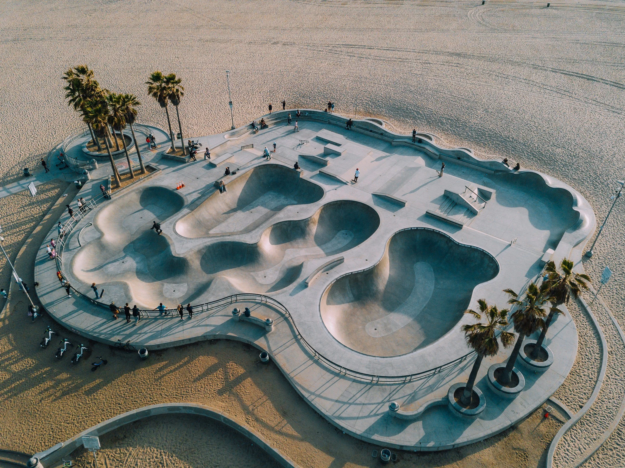 An aerial view of a skate park with various ramps and bowls, surrounded by a sandy area with a few palm trees and people walking and sitting nearby.