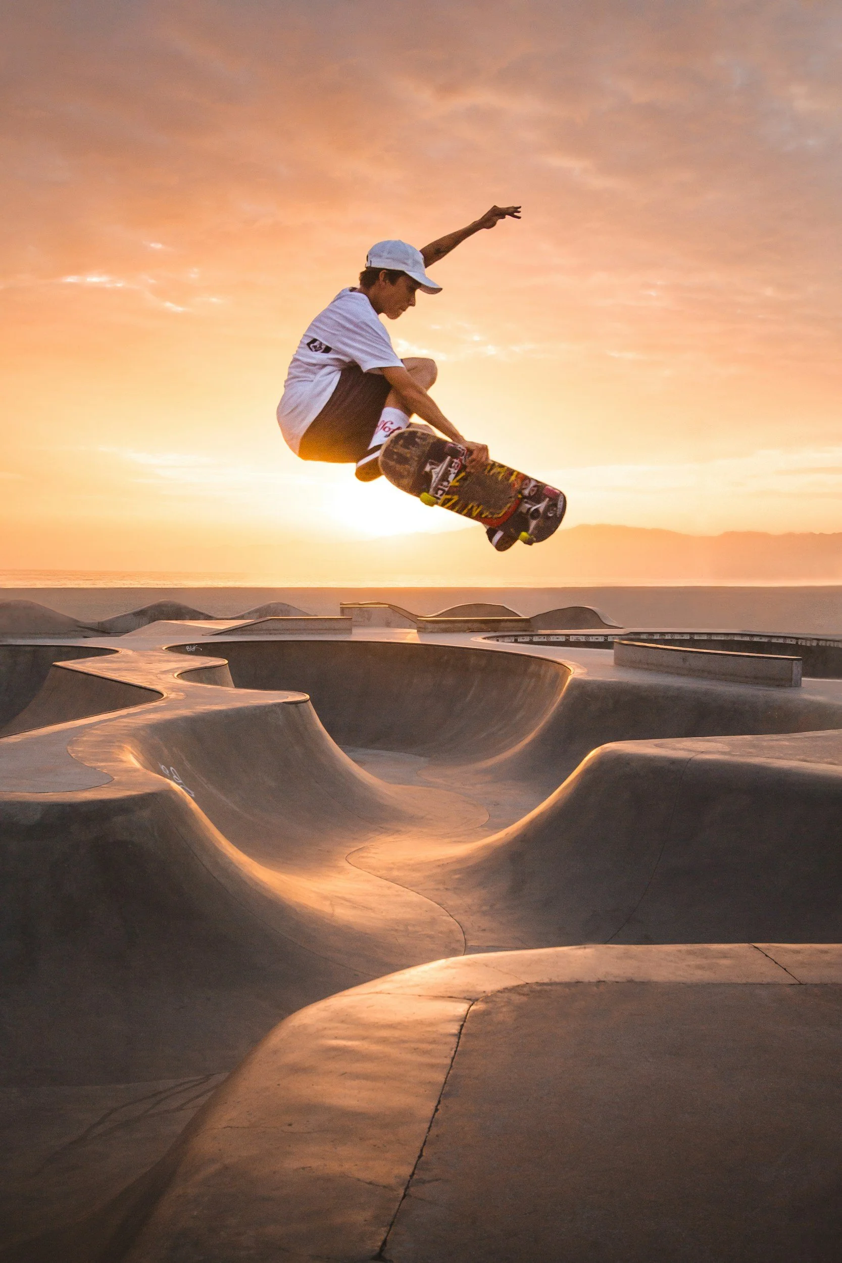 A young man wearing a white cap and t-shirt skateboarding at a skate park during sunset, with curving ramps and a colorful sky in the background.