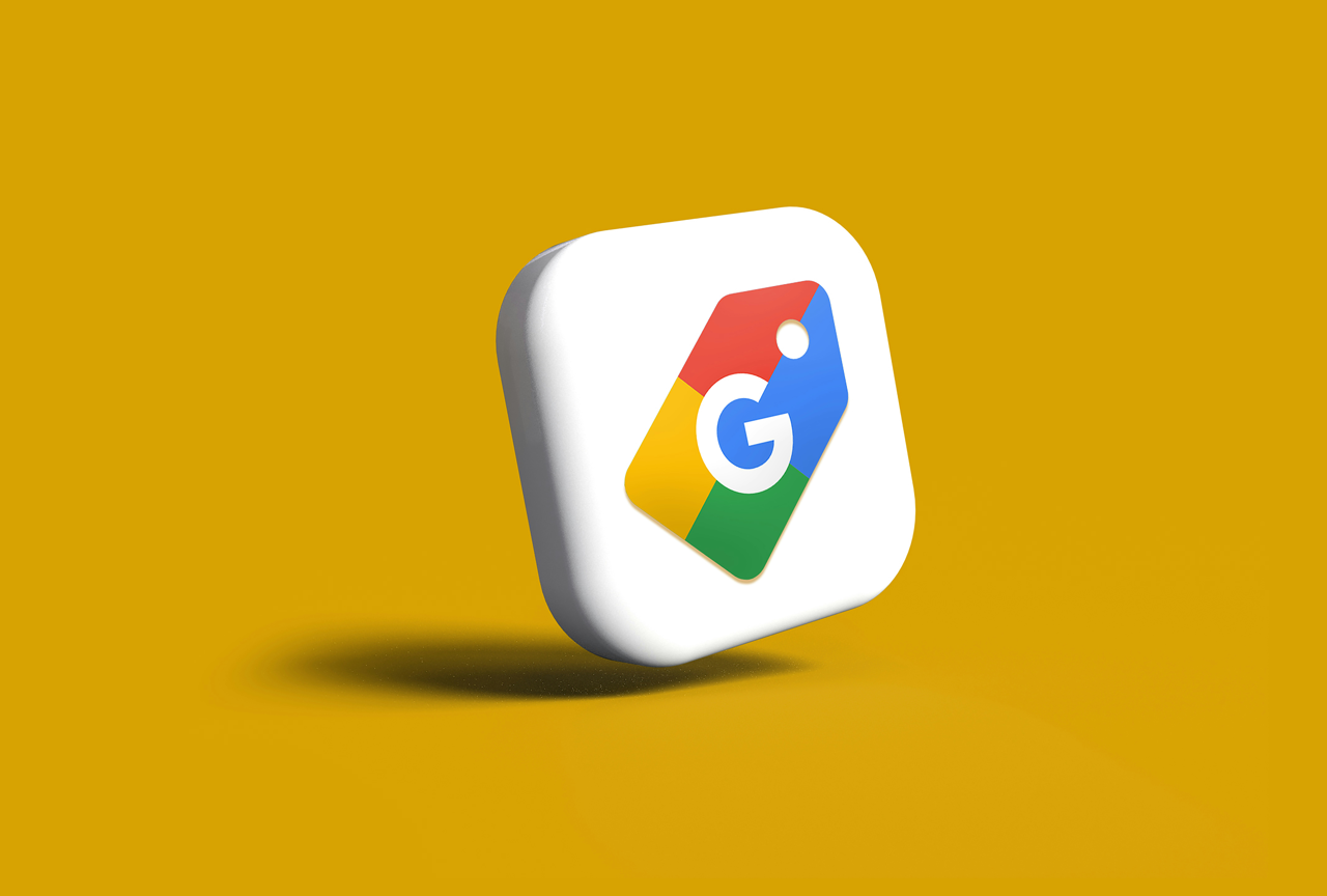 The image shows a three-dimensional rendering of an icon, which appears to be the logo for Google Merchant Center. The icon is a white, rounded square with a multicolored price tag or label design on it. The price tag is red, yellow, green, and blue,
