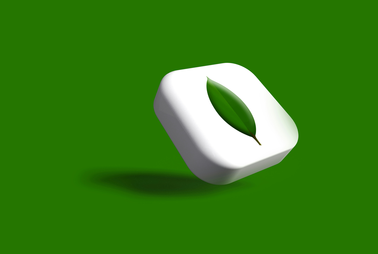The image shows a white, rounded, square-shaped 3D object floating against a dark green background. A single, deep green leaf is centered on the top face of the white object. The object casts a dark shadow underneath it.