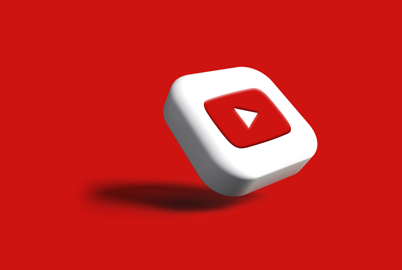 The image depicts a 3D rendering of the YouTube logo, which is a white square with rounded corners, elevated and angled against a plain red background. A red square with a white, right-pointing play button triangle is visible on the top surface of th