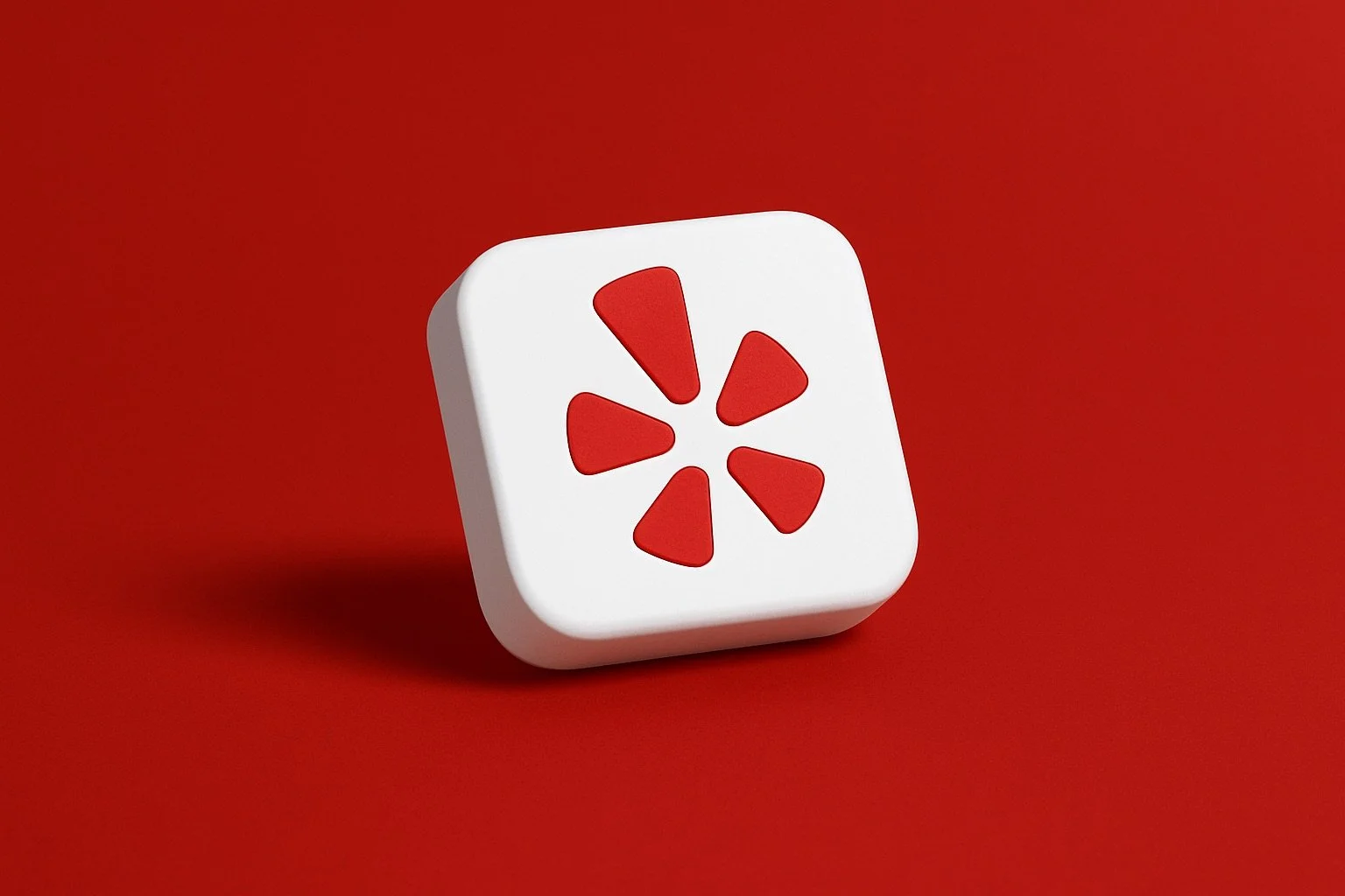 The image shows a three-dimensional, white, rounded square with the red Yelp logo embedded in its center, all set against a solid red background. The Yelp logo resembles a five-pointed star or an asterisk, where each point is a separate, rounded tria