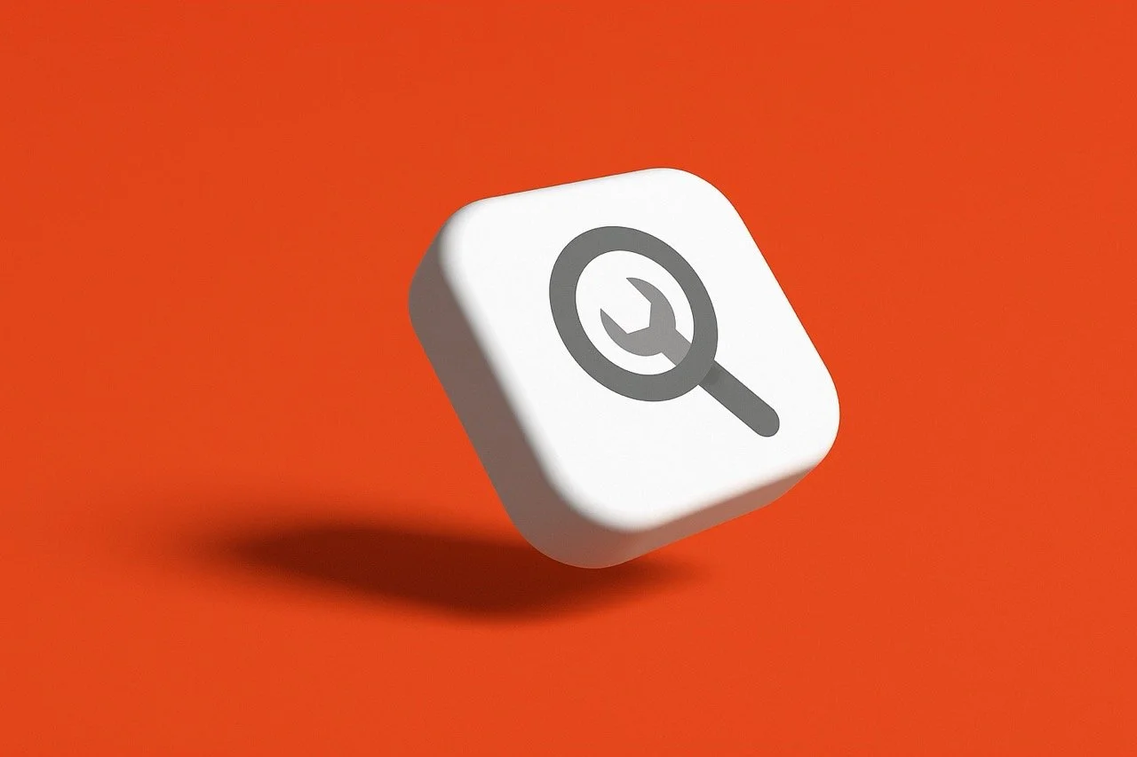 The image depicts a white, three-dimensional, slightly rounded cube against a solid, bright orange background. On the top surface of the cube, there is a gray icon that combines a magnifying glass and a wrench. The cube appears to be floating or susp