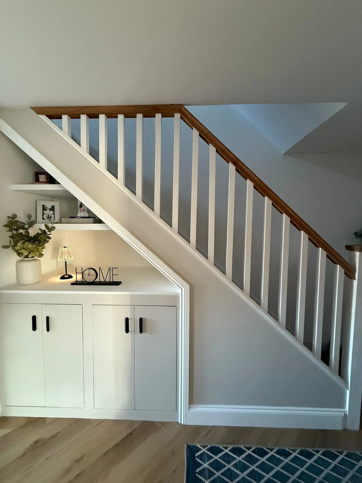 Interior of a home featuring a staircase with white risers and a wooden handrail. Beneath the staircase, there is a white cabinet with black handles, a small table lamp, a decorative 'HOME' sign, a 'LOVE' picture frame, and a vase with green flowers. Part of a blue patterned rug is visible on the wooden floor.